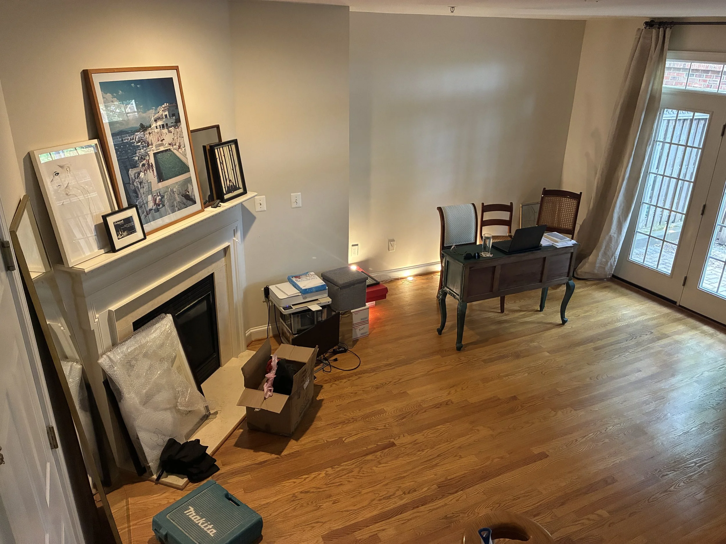 A living room in the midst of a move, showing the messy middle of decorating with unpacked boxes, framed artwork leaning against a white mantle, and a temporary home office setup.