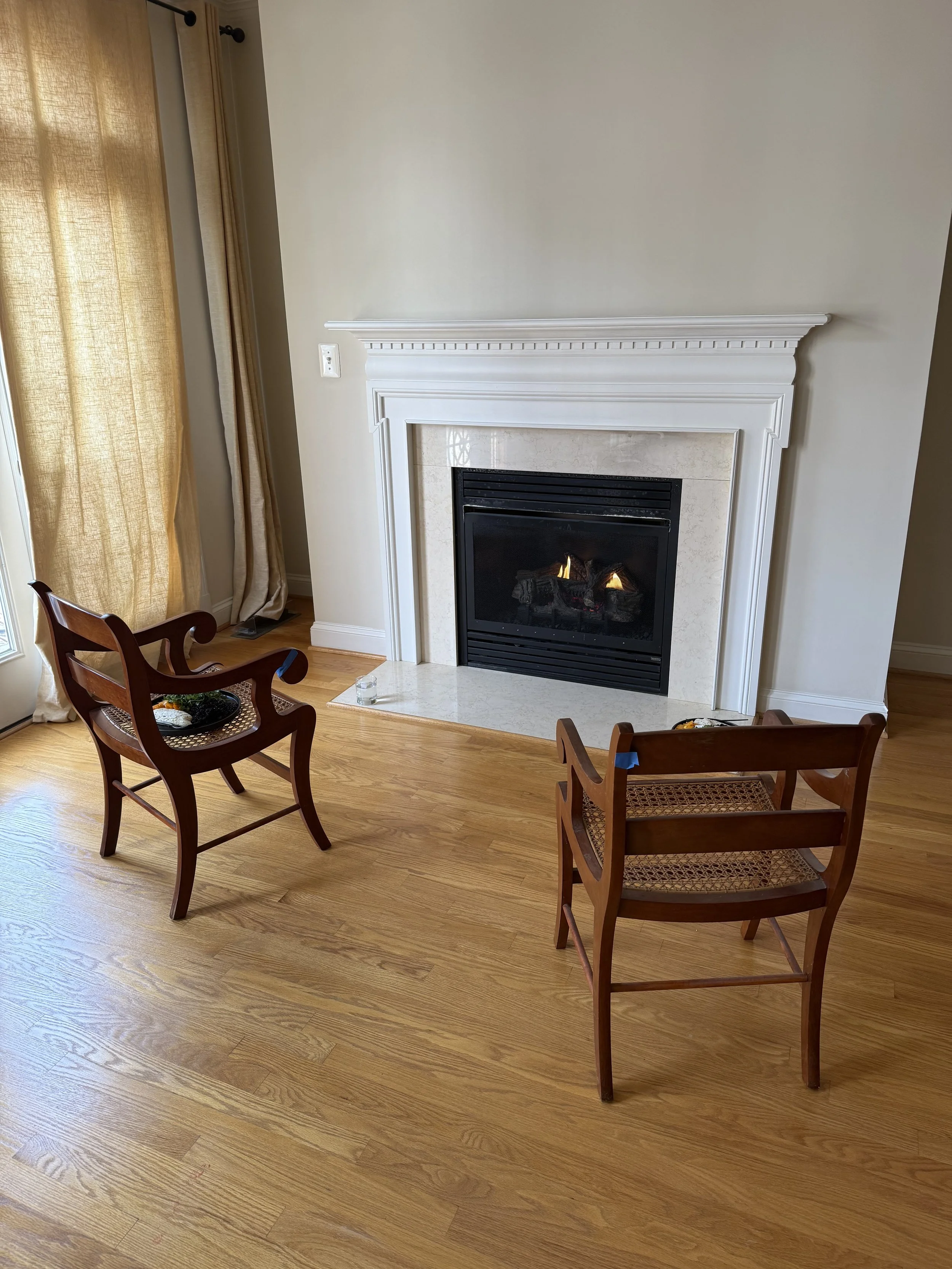 A curated rental living room in McLean Virginia featuring a white fireplace, vintage wooden chairs, and neutral curtains.