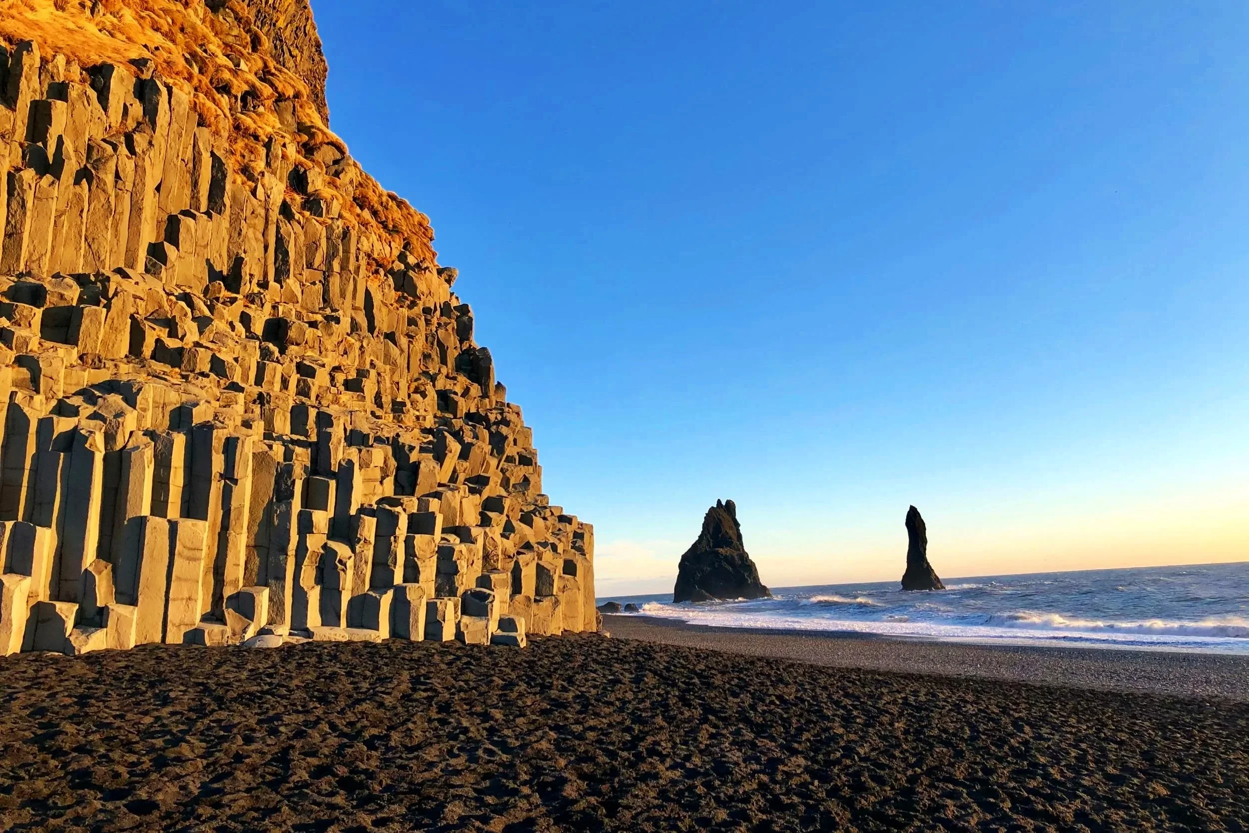 A black sand beach lined with tall, geometric basalt rock columns next to the ocean with sea stacks in the distance, featuring the word Adventure.