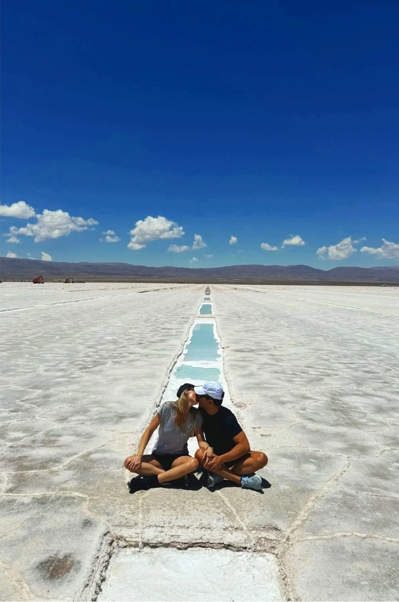 A couple sitting cross-legged and kissing on an expansive white salt flat under a bright blue sky, below the word Family.