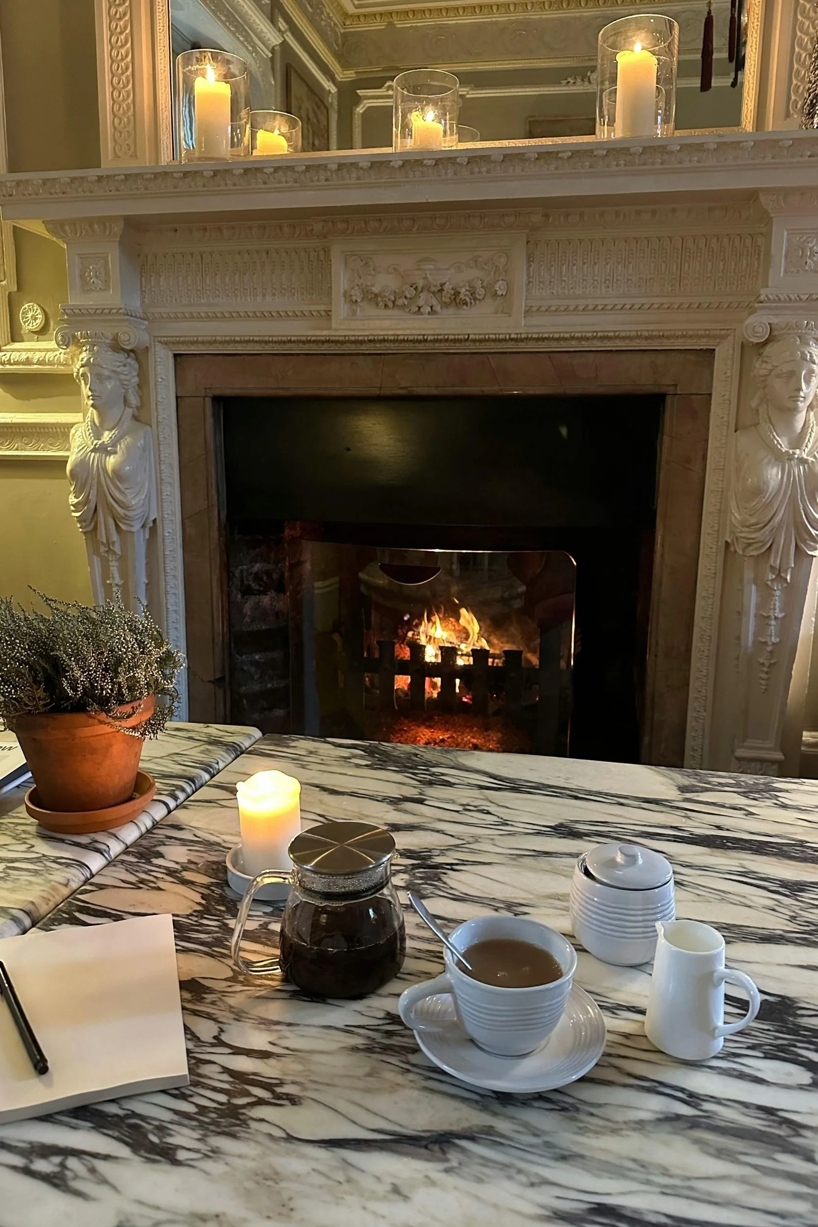A marble table set with a teapot, teacup, notebook, and a candle in front of an ornate white fireplace with a lit fire and a mirror reflecting candles on the mantel.