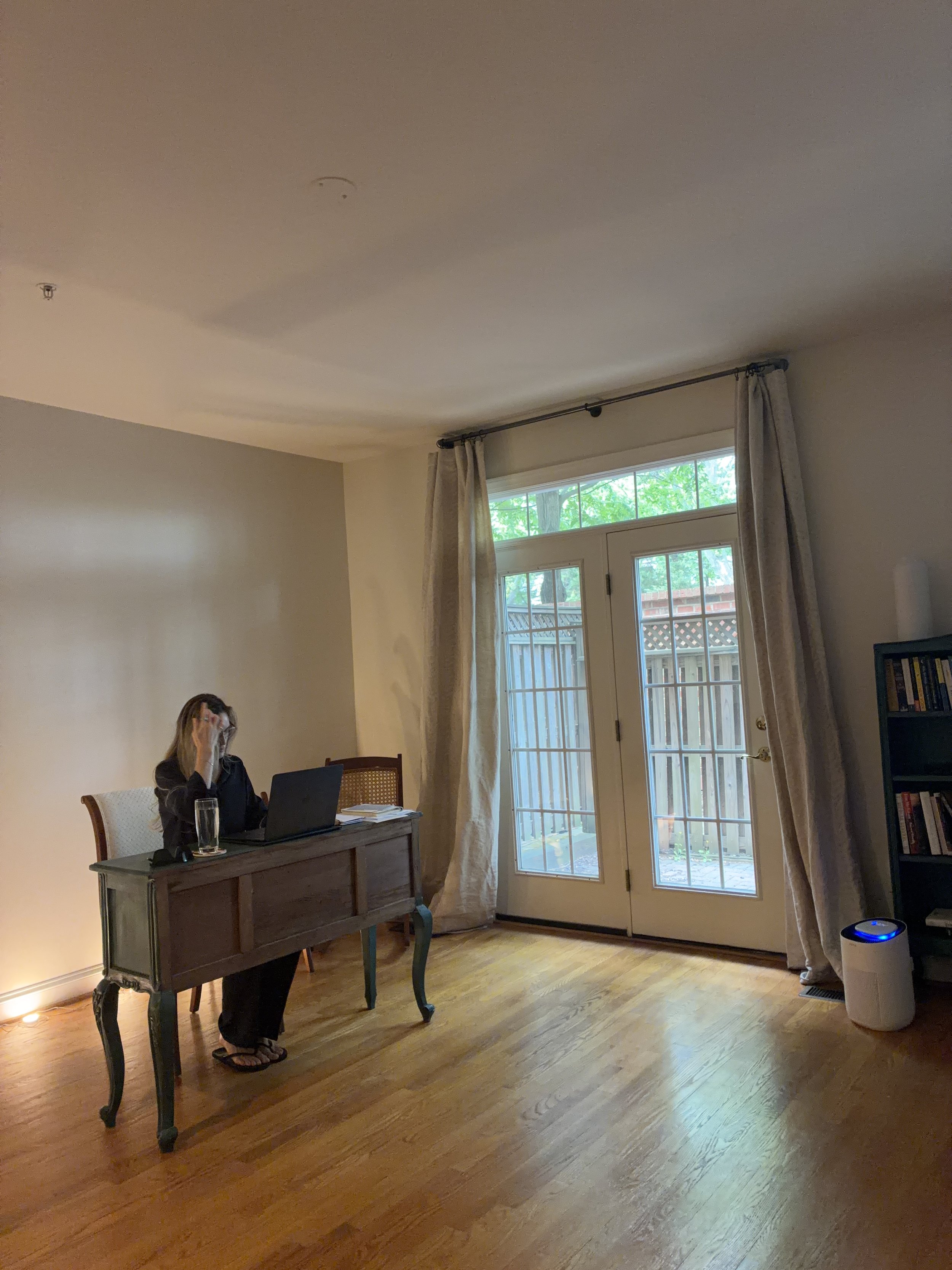 A woman working on a laptop at a vintage wooden desk in a sparsely furnished, sunlit room with French doors, illustrating the transitional reality of working remotely during a move.