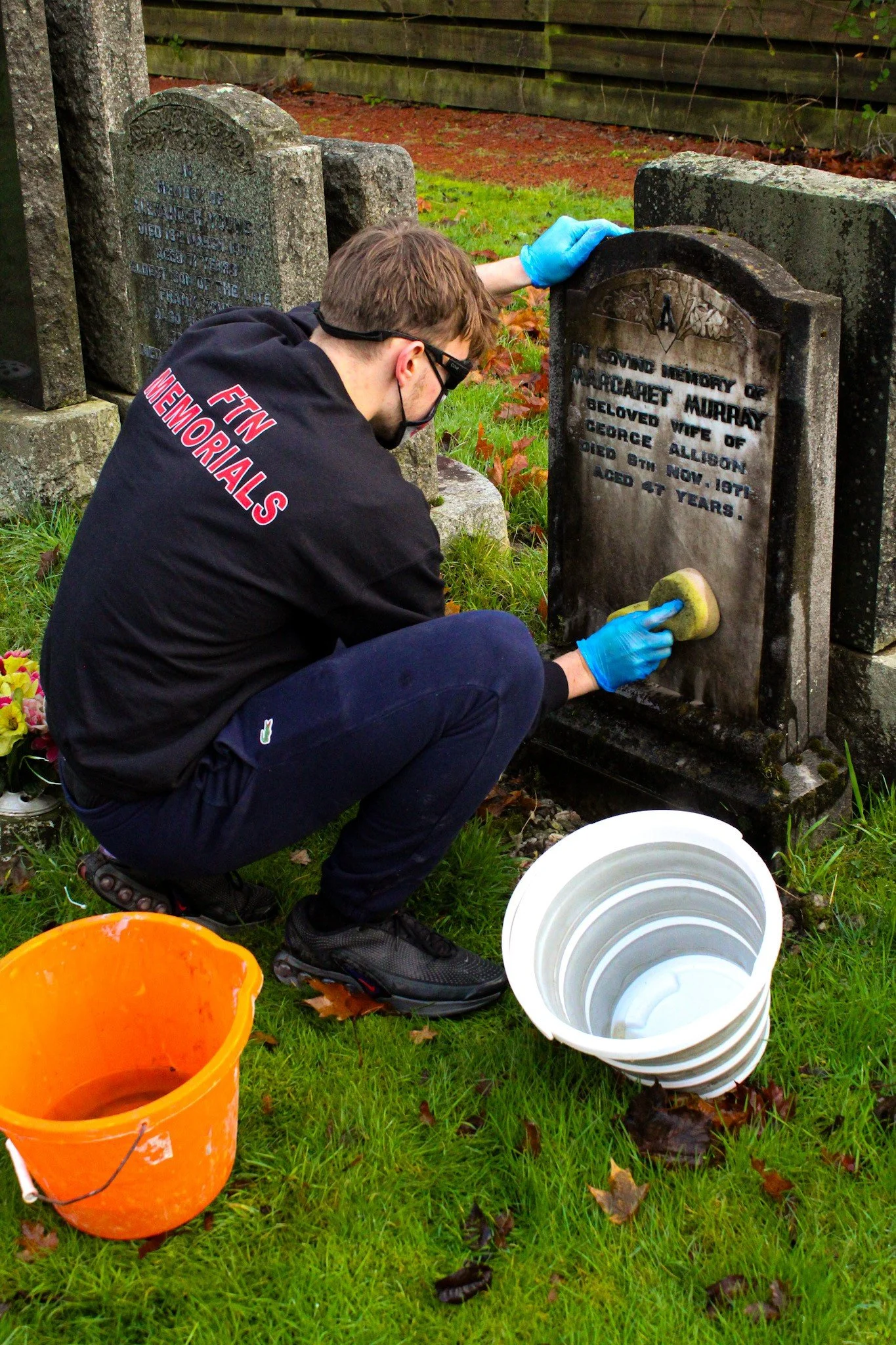 A person wearing a black fleece, glasses, and gloves is cleaning a black marble tombstone with a sponge, in a cemetery. An orange bucket and white buckets are placed on the grass nearby.