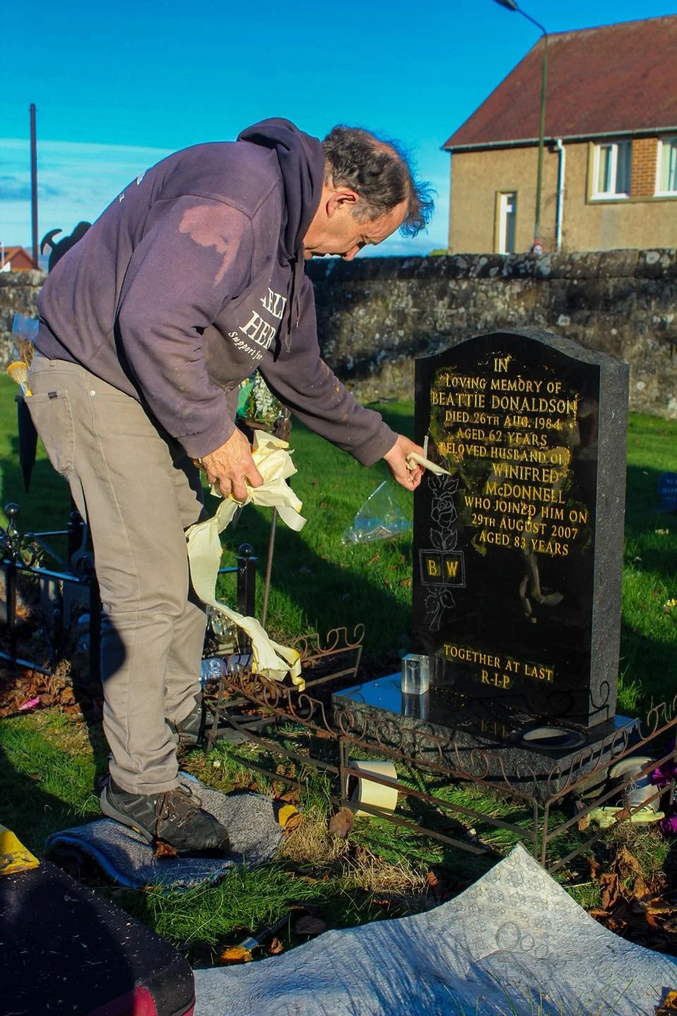 A man kneels at a grave, cleaning and maintaining the headstone with a brush, in a cemetery on a sunny day.