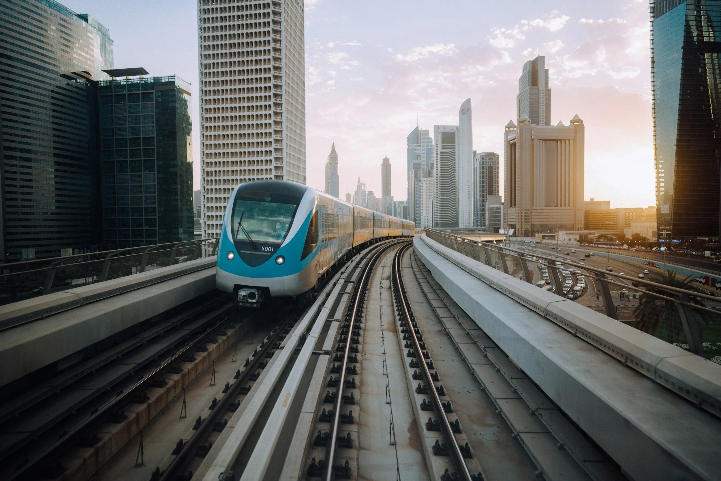 A modern train traveling on elevated tracks through a city with tall skyscrapers during sunset.