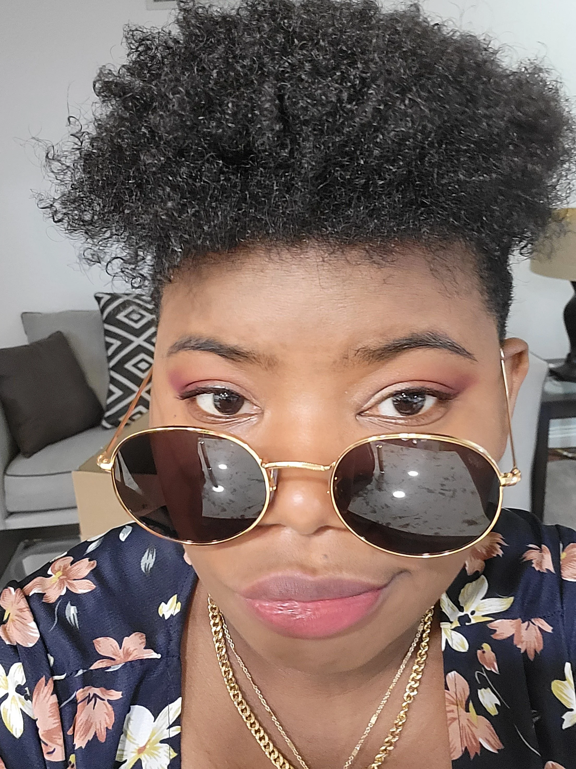 Close-up photo of a woman wearing aviator sunglasses, a floral blouse, and gold jewelry. She has short, curly hair and is indoors, with a sofa and patterned pillow visible in the background.