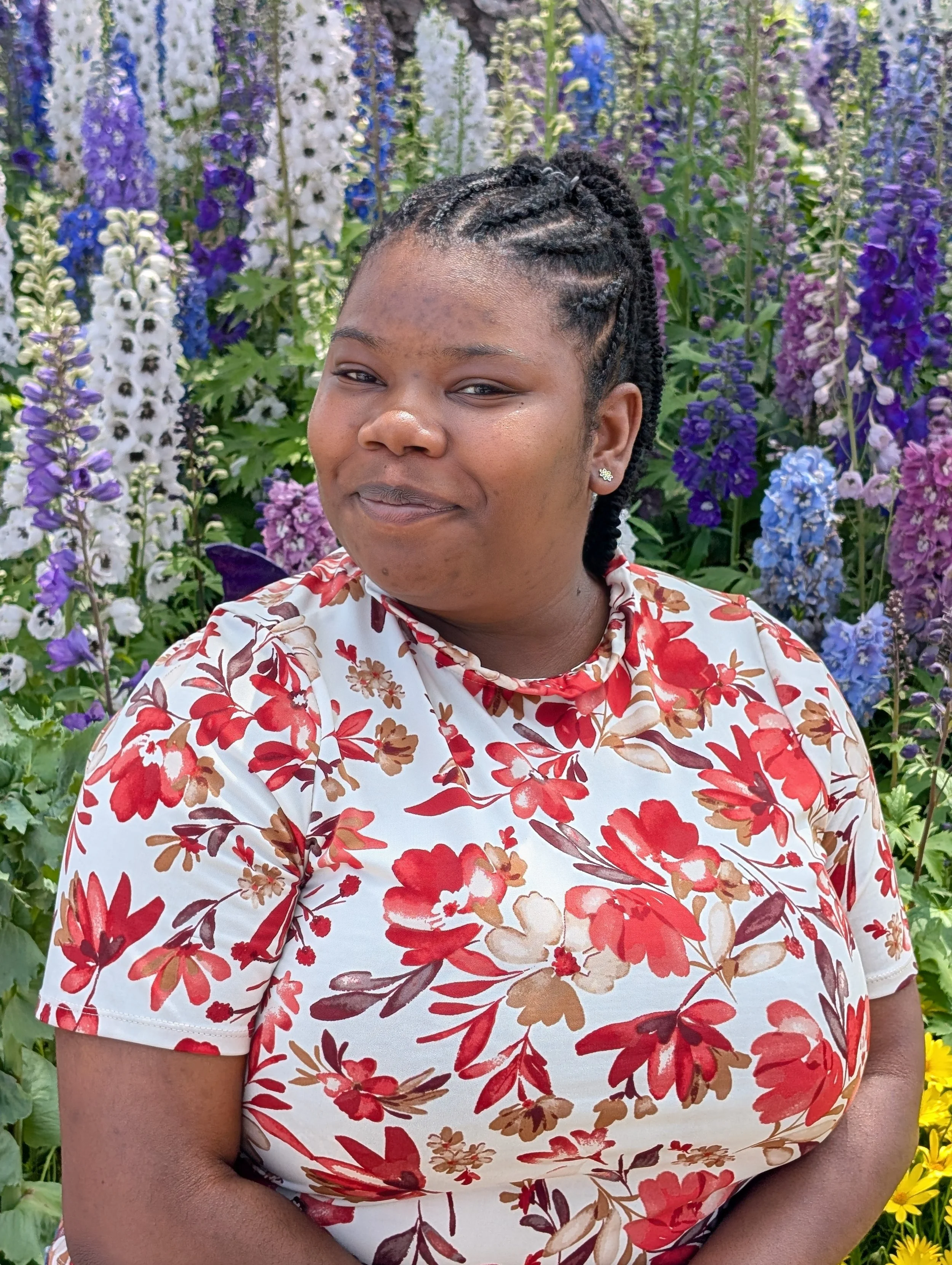 Portrait of a woman with braided hair and earrings, wearing a floral shirt, standing in front of a colorful flower garden with tall purple, white, and blue flowers.