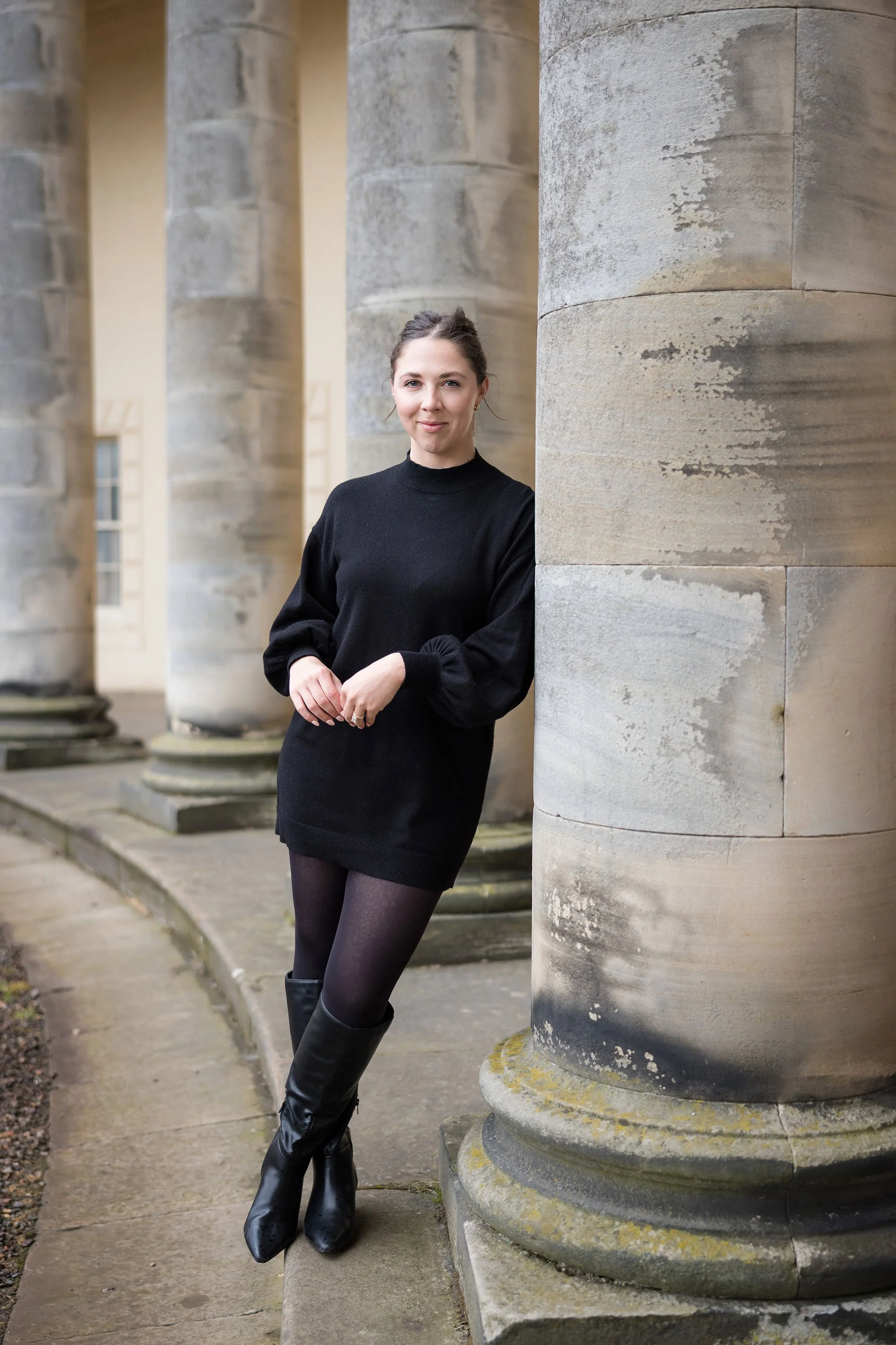 A woman in a black sweater dress and black boots leaning against a large stone column outside a historic building.