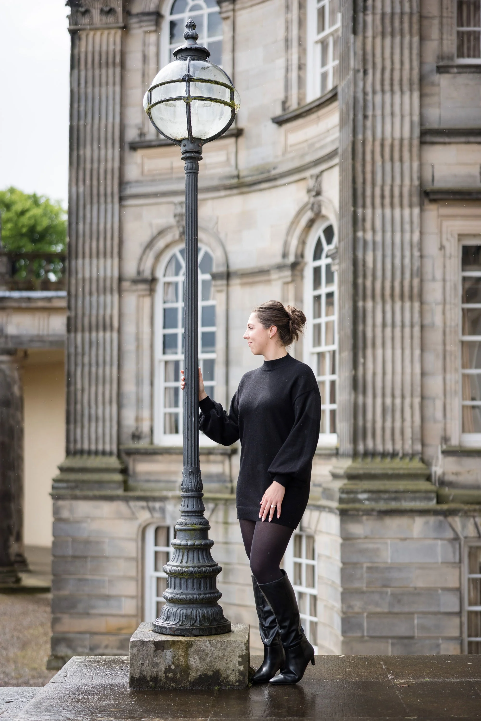A woman in black dress and boots standing next to an old-fashioned street lamp outside a historic stone building with large arched windows.