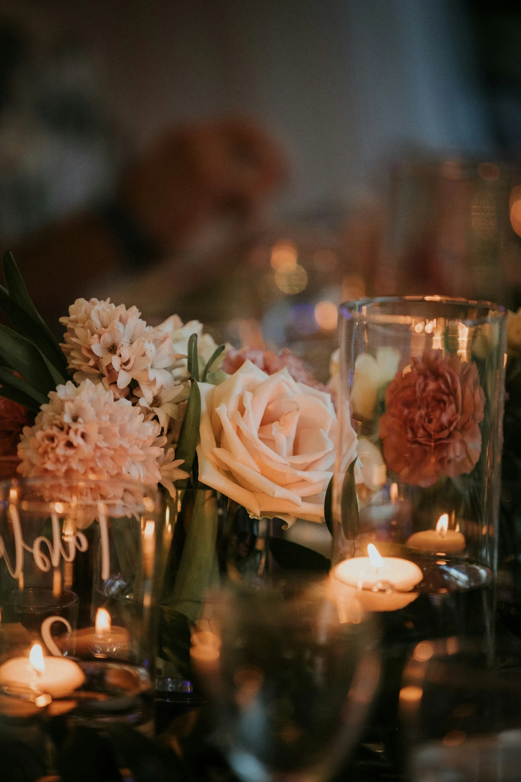 A floral centerpiece with white roses, light pink flowers, and greenery, surrounded by candles in glass holders, on a dimly lit table.