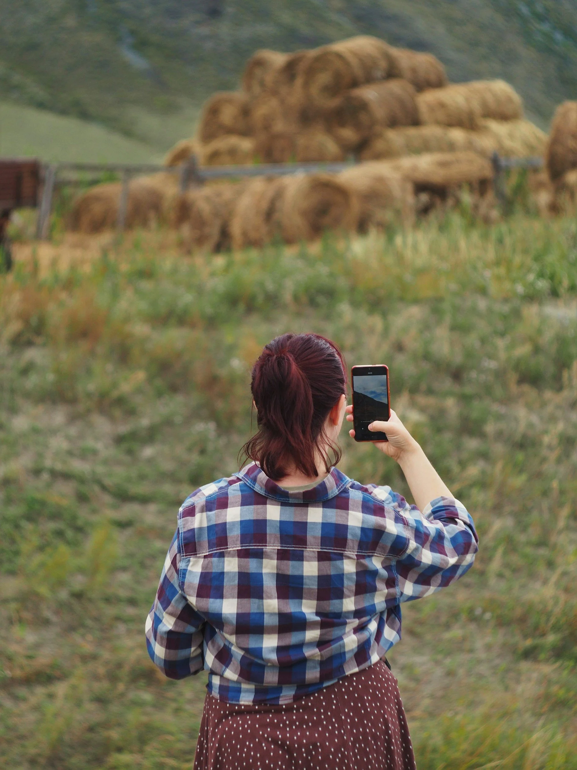 A woman with red hair, wearing a plaid shirt and holding a smartphone, taking a photo of a haystack in a rural area.
