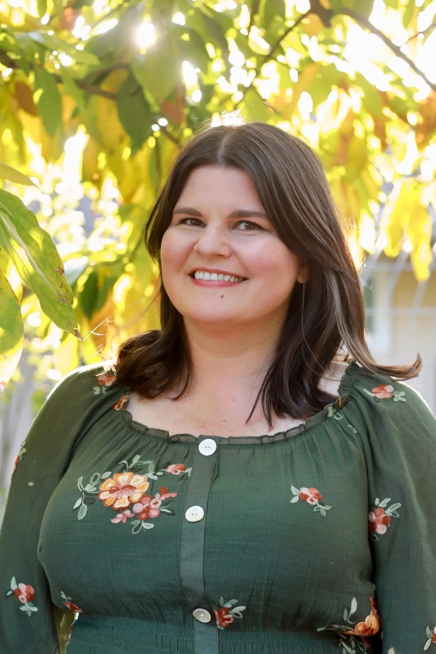 A woman with dark brown hair, smiling, standing outdoors among green leaves with sunlight shining through.