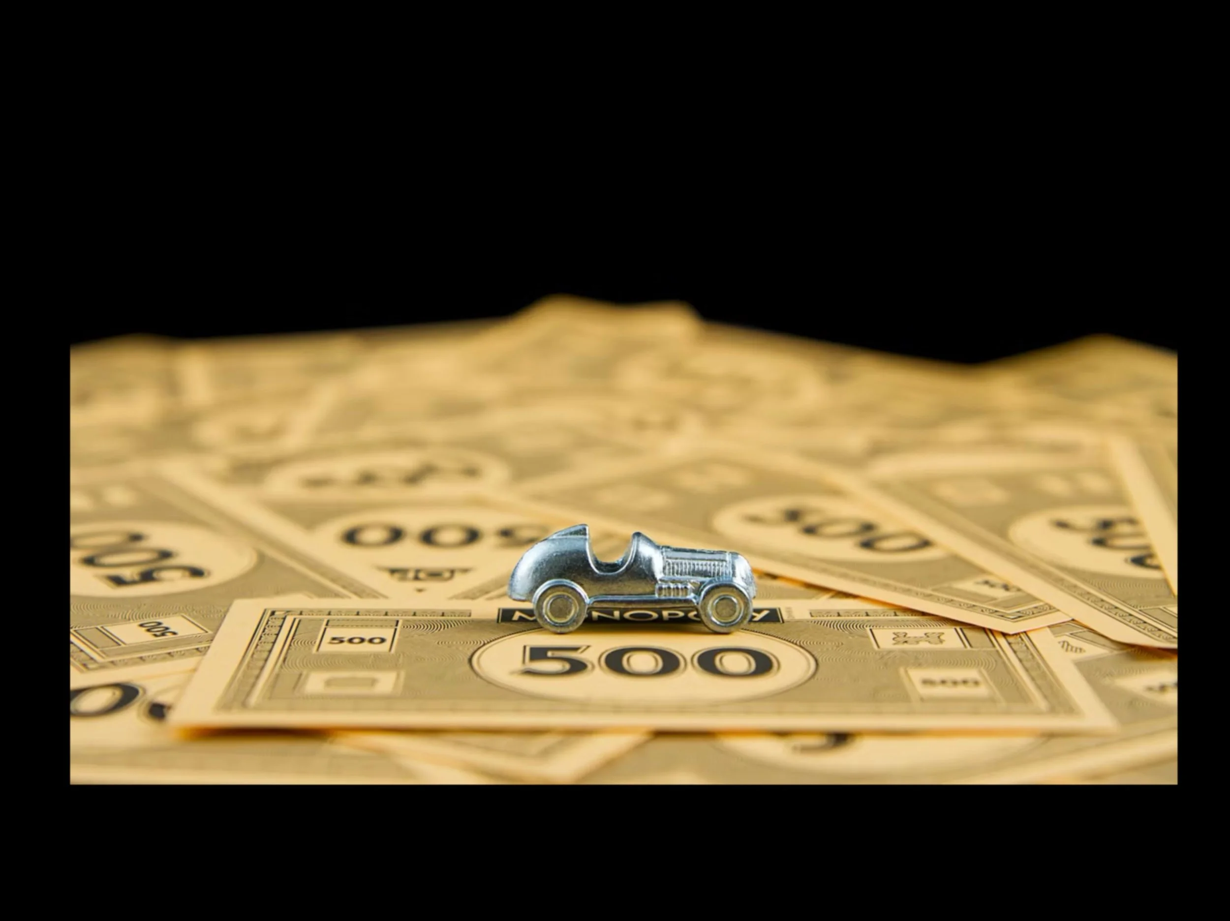 Tiny metal car toy sitting on scattered US $500 bills with a black background.
