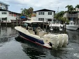 A beige motorboat with twin outboard engines in a harbor, surrounded by other boats and houses on stilts.