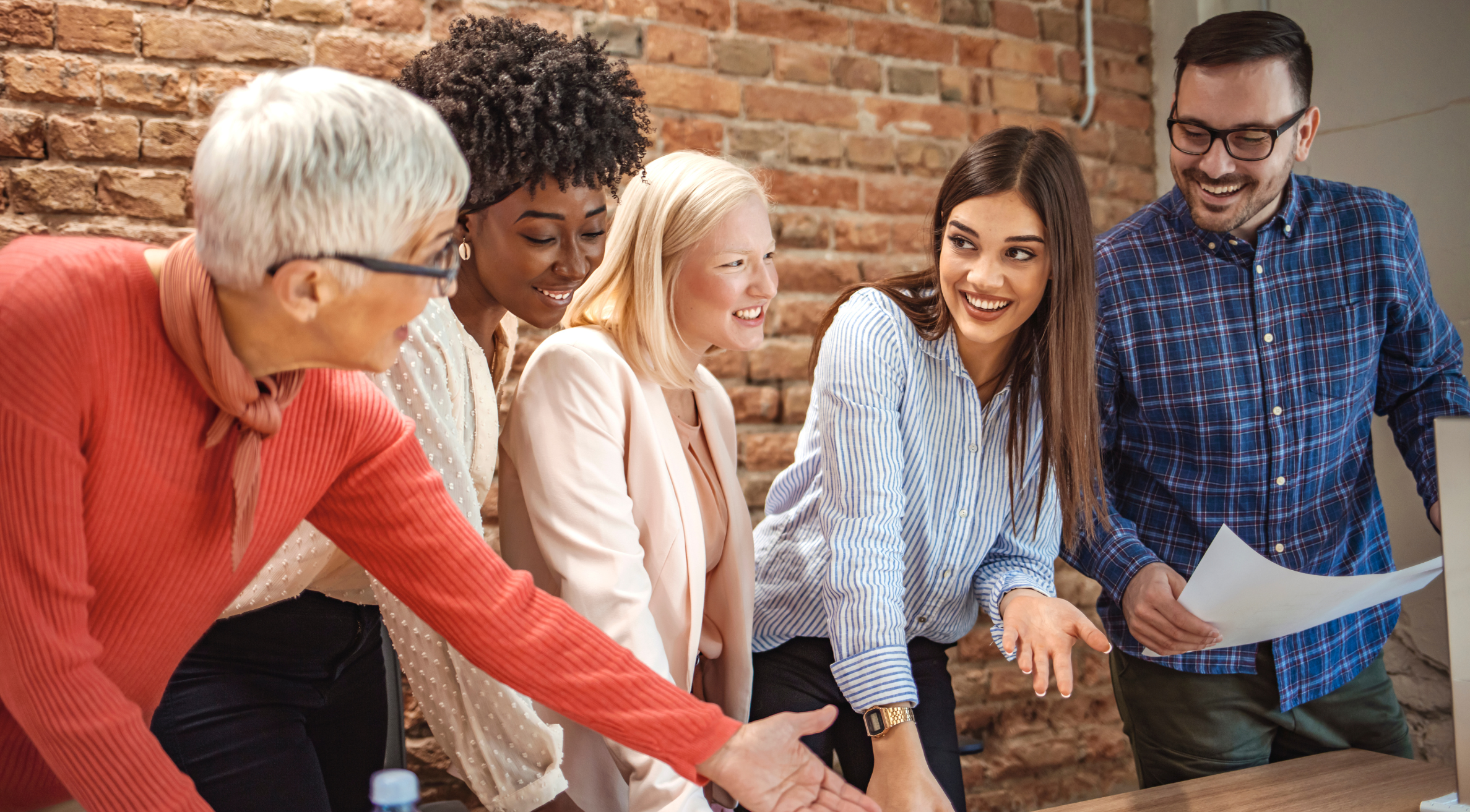 Group of five diverse coworkers gathered around a table, smiling, and engaged in a discussion in an office with exposed brick walls.
