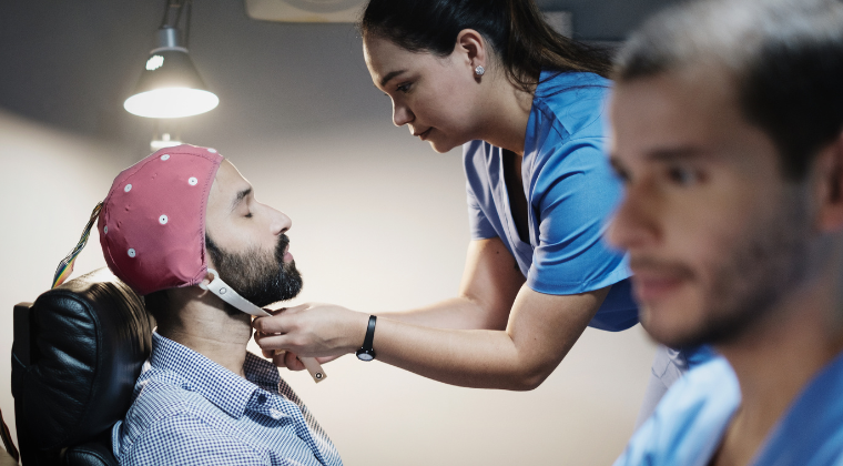 A healthcare professional adjusting a sleep study device on a male patient's head in a clinical setting, with another healthcare worker in the background.
