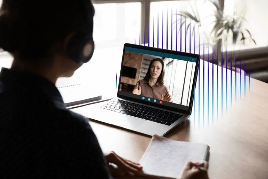 Person wearing headphones participating in a video call on a laptop, with notes and a notebook on the table, in a well-lit room with a window and plant in the background.