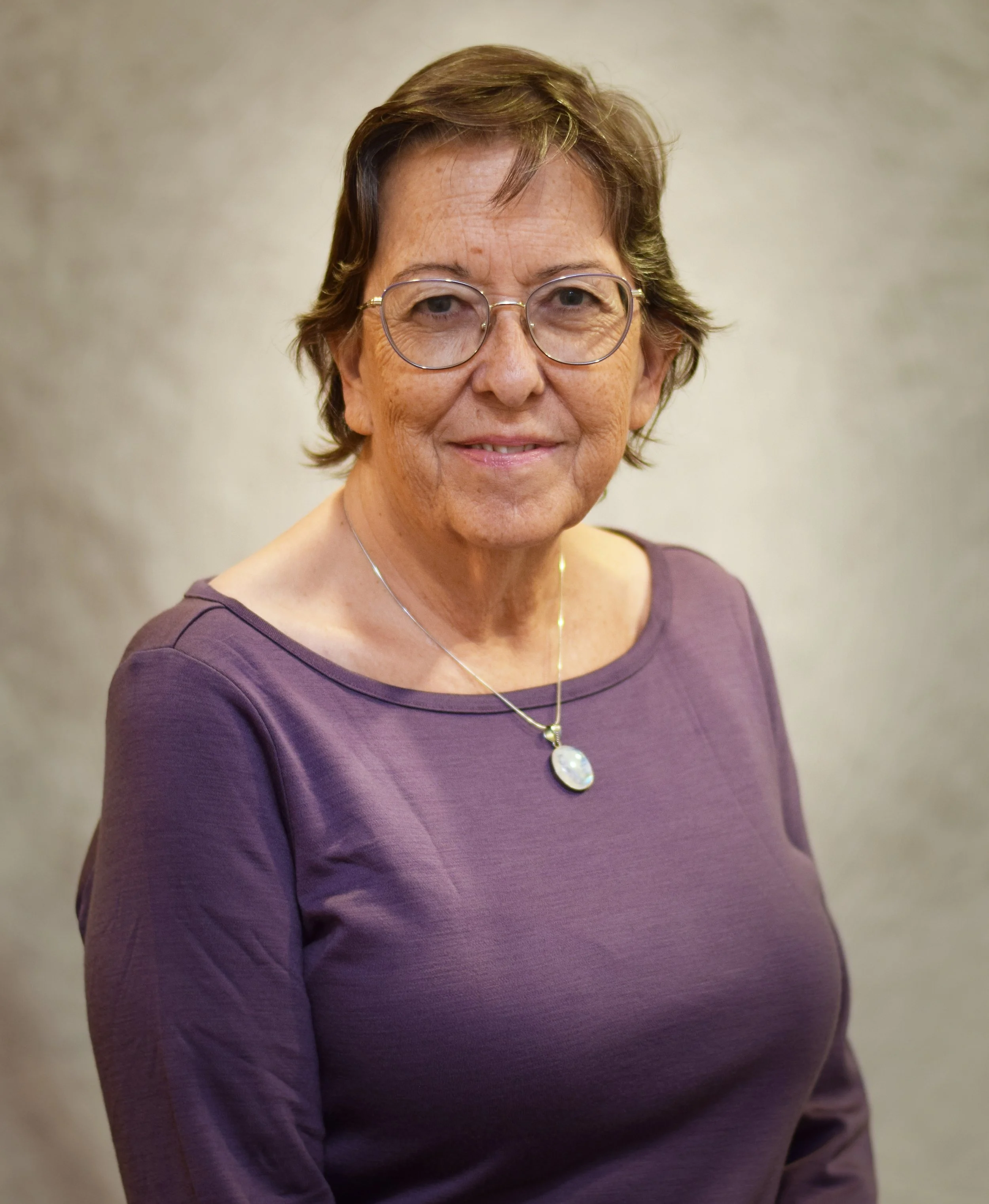 A middle-aged woman with short brown hair, glasses, and a purple top, smiling at the camera against a beige background.