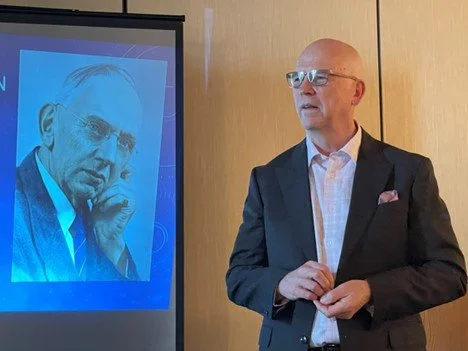 A bald man with glasses wearing a dark suit and white shirt, standing in front of a wooden wall, giving a presentation.