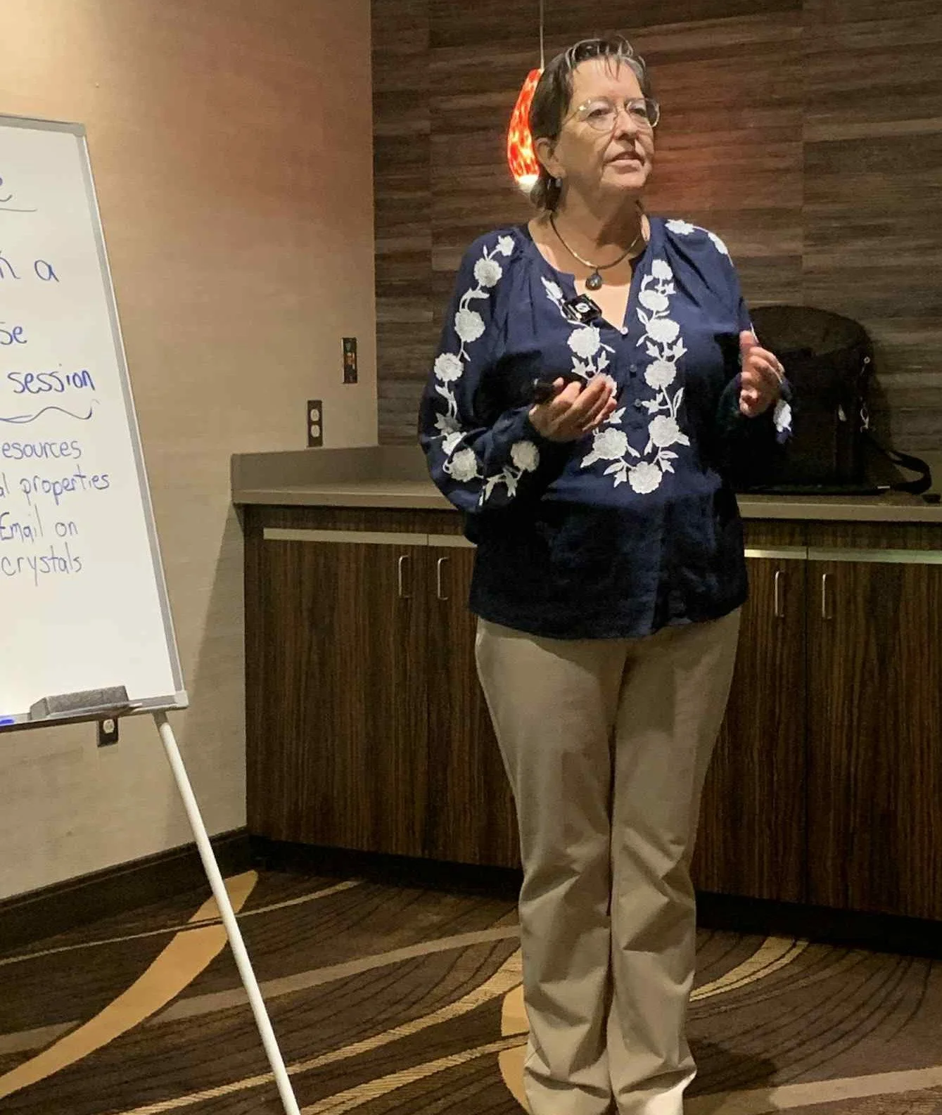 A woman giving a presentation in a conference room, standing next to a whiteboard with notes on it, holding a remote control in her hand.