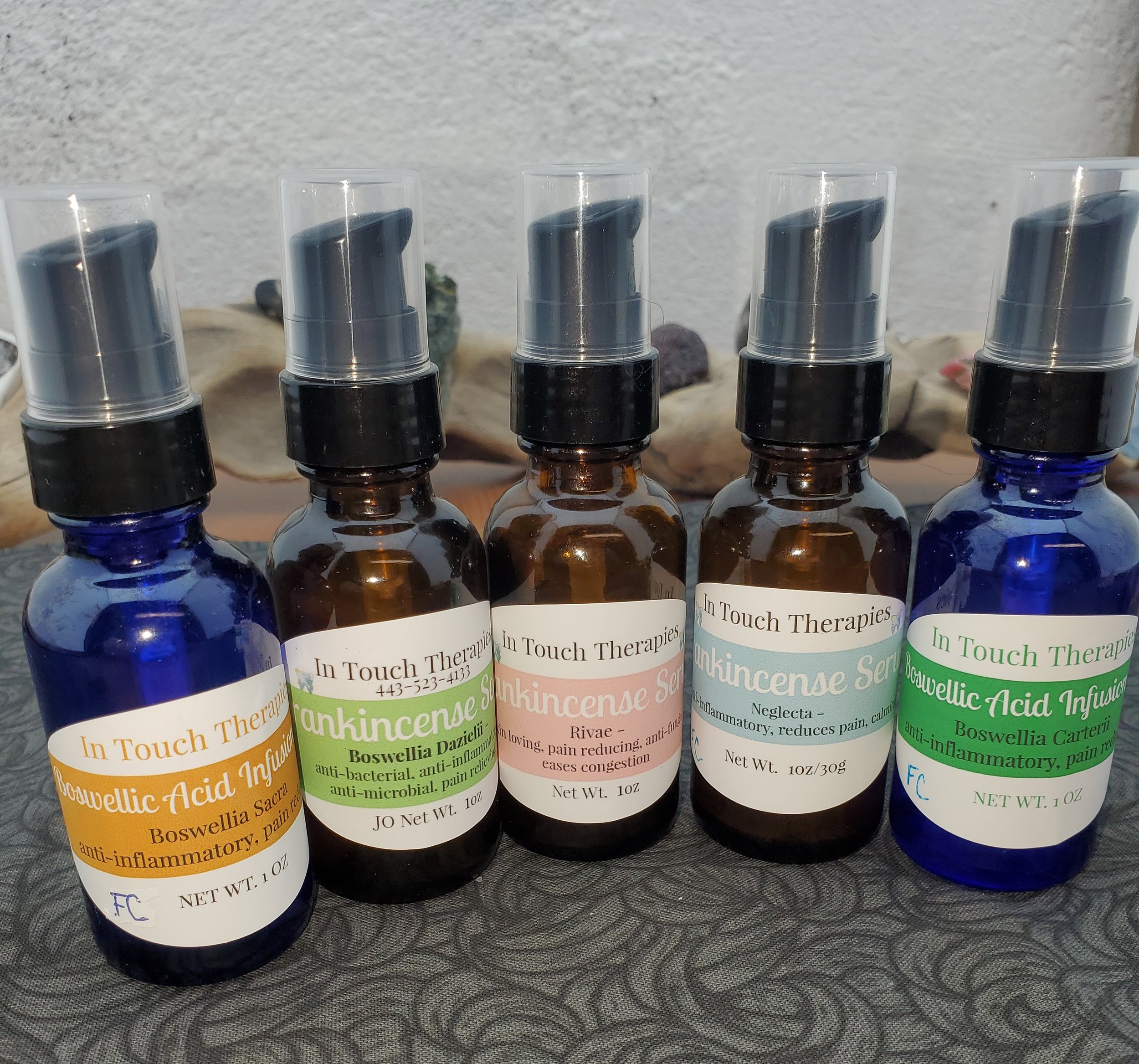 Five small bottles with spray tops labeled 'In Touch Therapies' containing Boswellia extracts with different properties, arranged on a textured surface with seashells in the background.