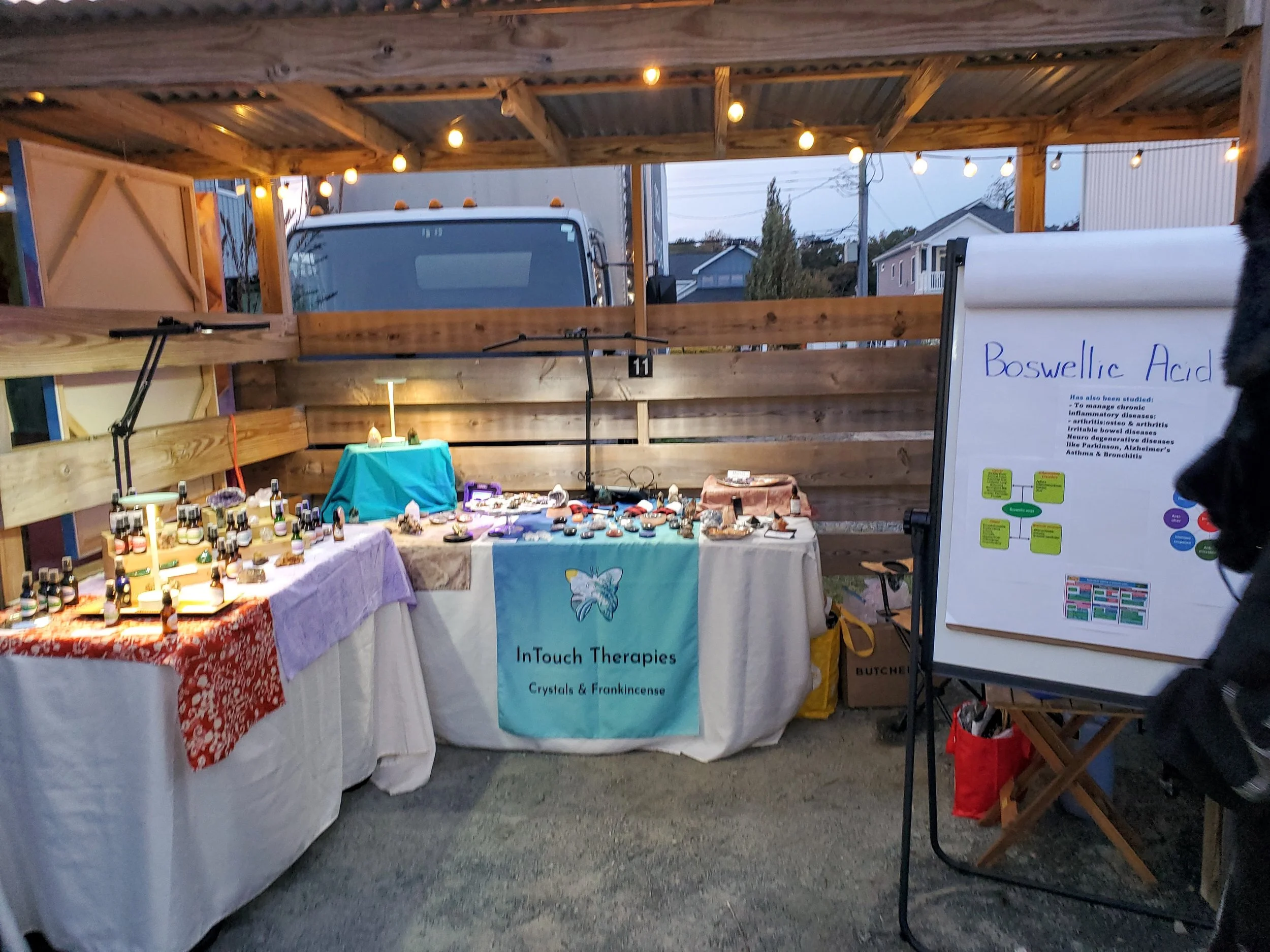 An outdoor market stall selling essential oils and crystals under string lights, with a wooden canopy, a truck in the background, and a large whiteboard with information about Boswellic Acid to the right.