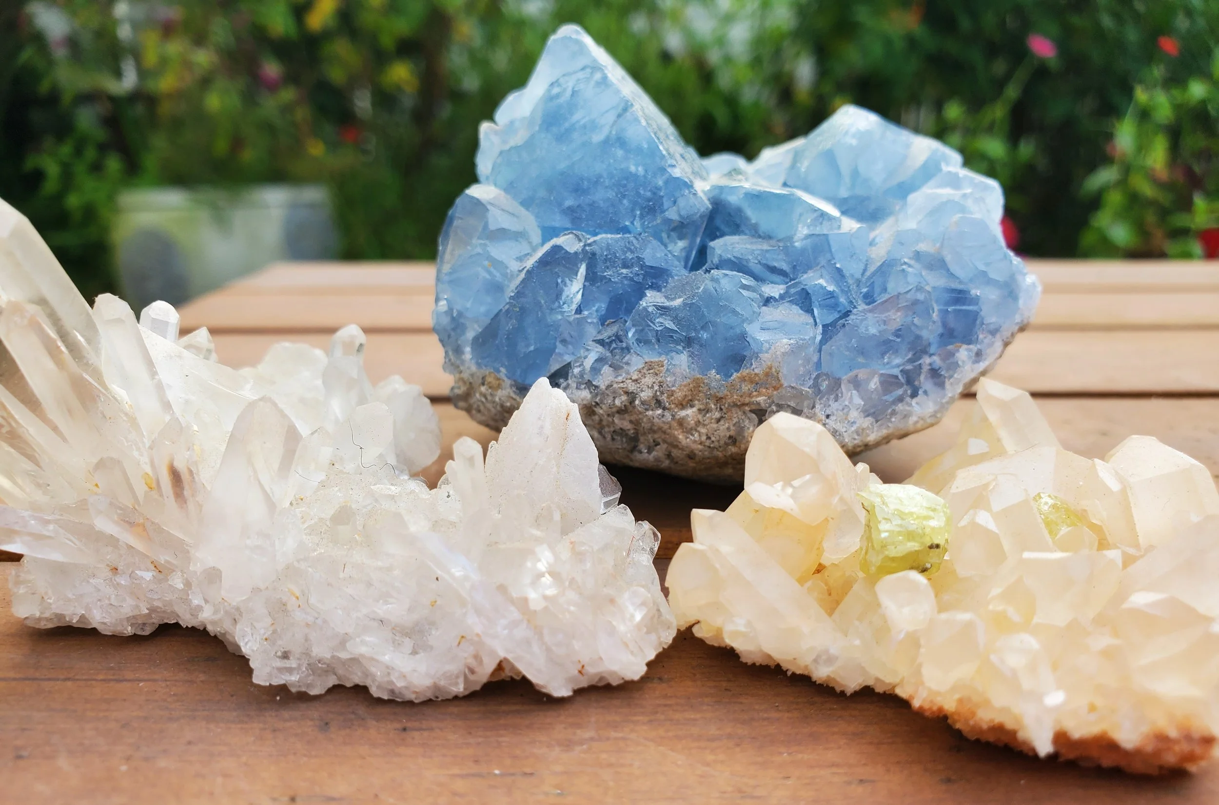 Three large crystalline mineral specimens, including a blue mineral with a rough base and transparent crystals, one white mineral with a cluster of sharp, white crystals, and a pale yellow mineral with small green inclusions, placed on a wooden surface outdoors.