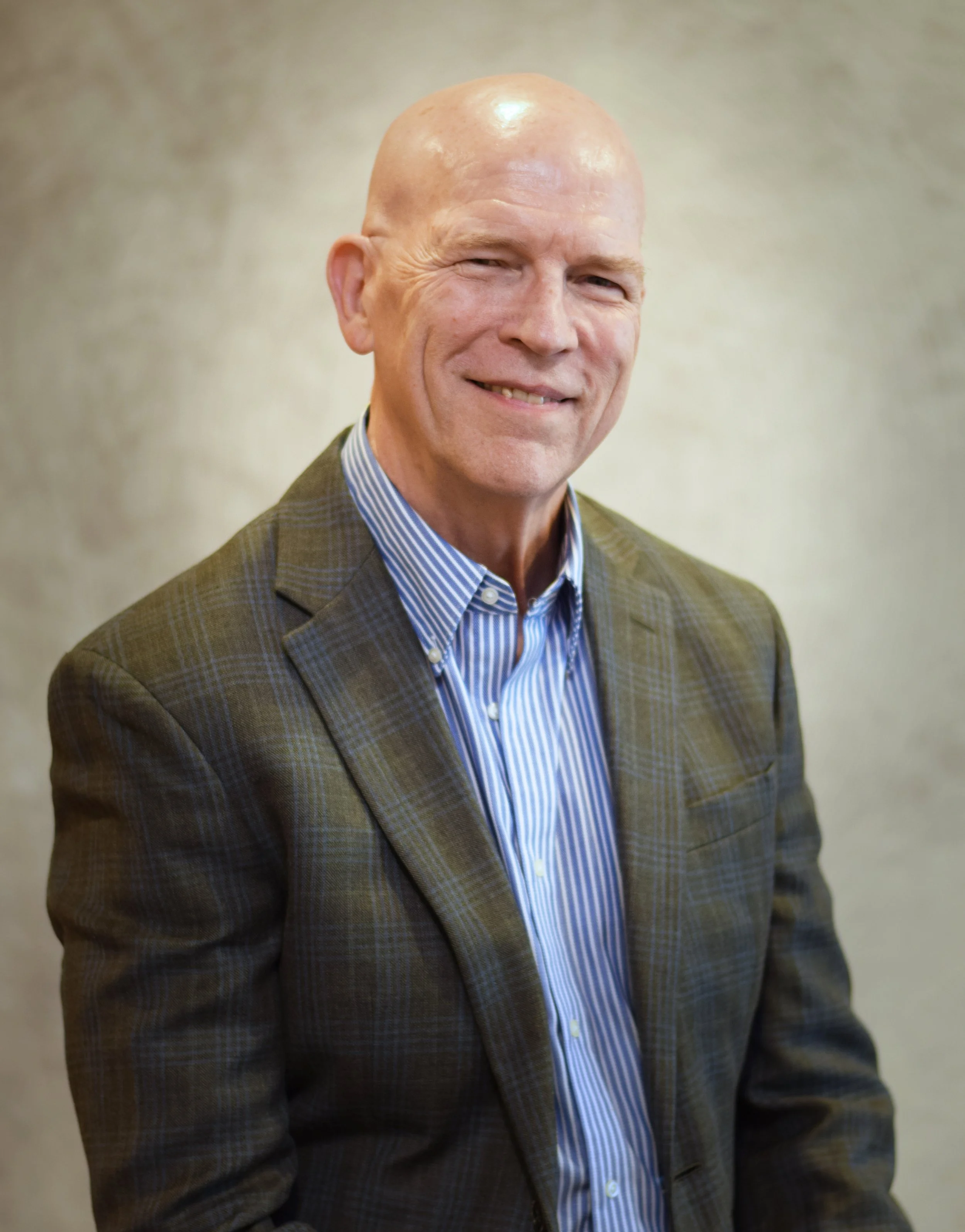 A middle-aged man with a bald head wearing a plaid blazer and a striped blue shirt, smiling at the camera against a neutral background.