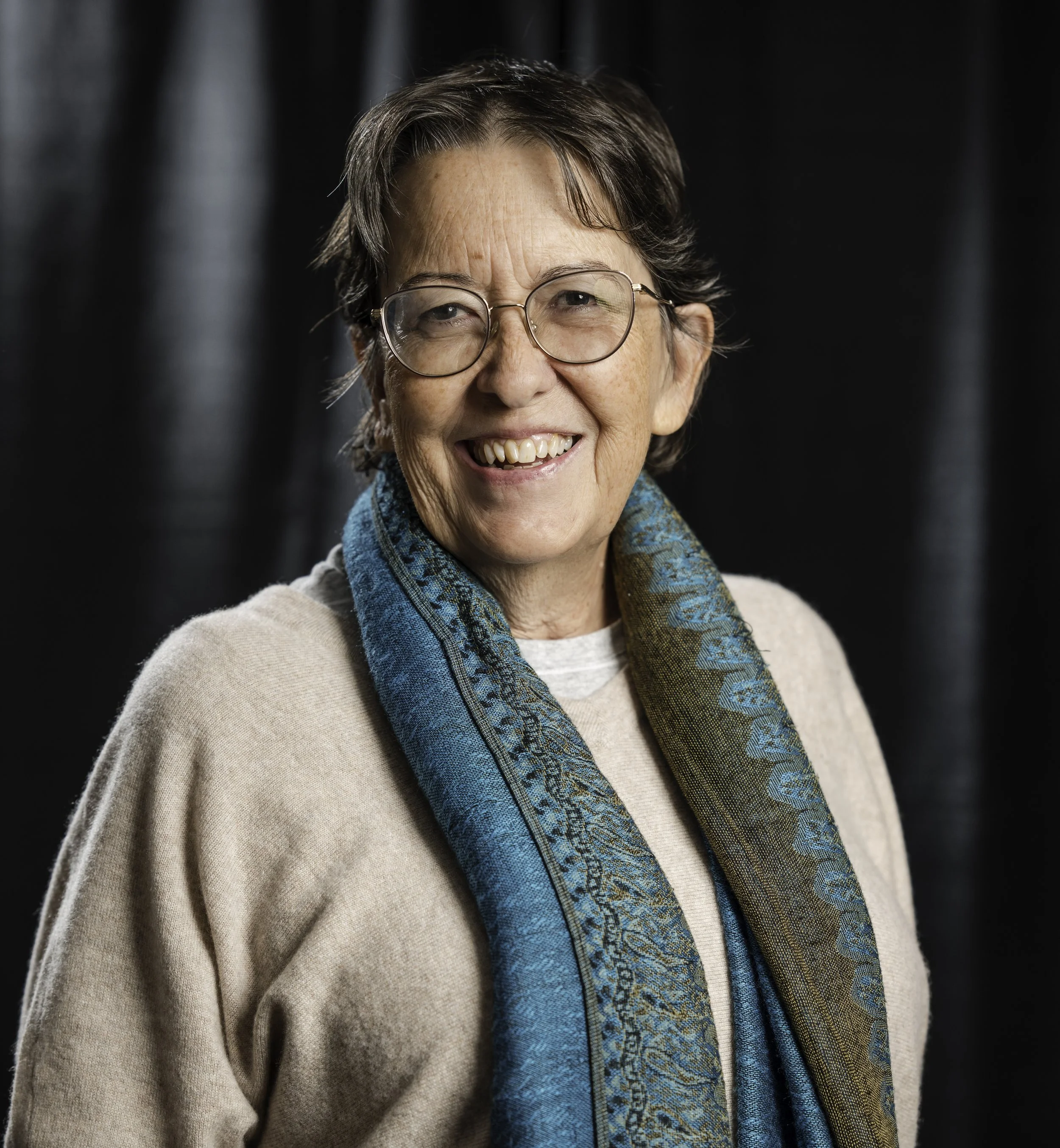 A smiling elderly woman with glasses, short gray hair, wearing a beige sweater and a blue patterned scarf, standing against a dark background.