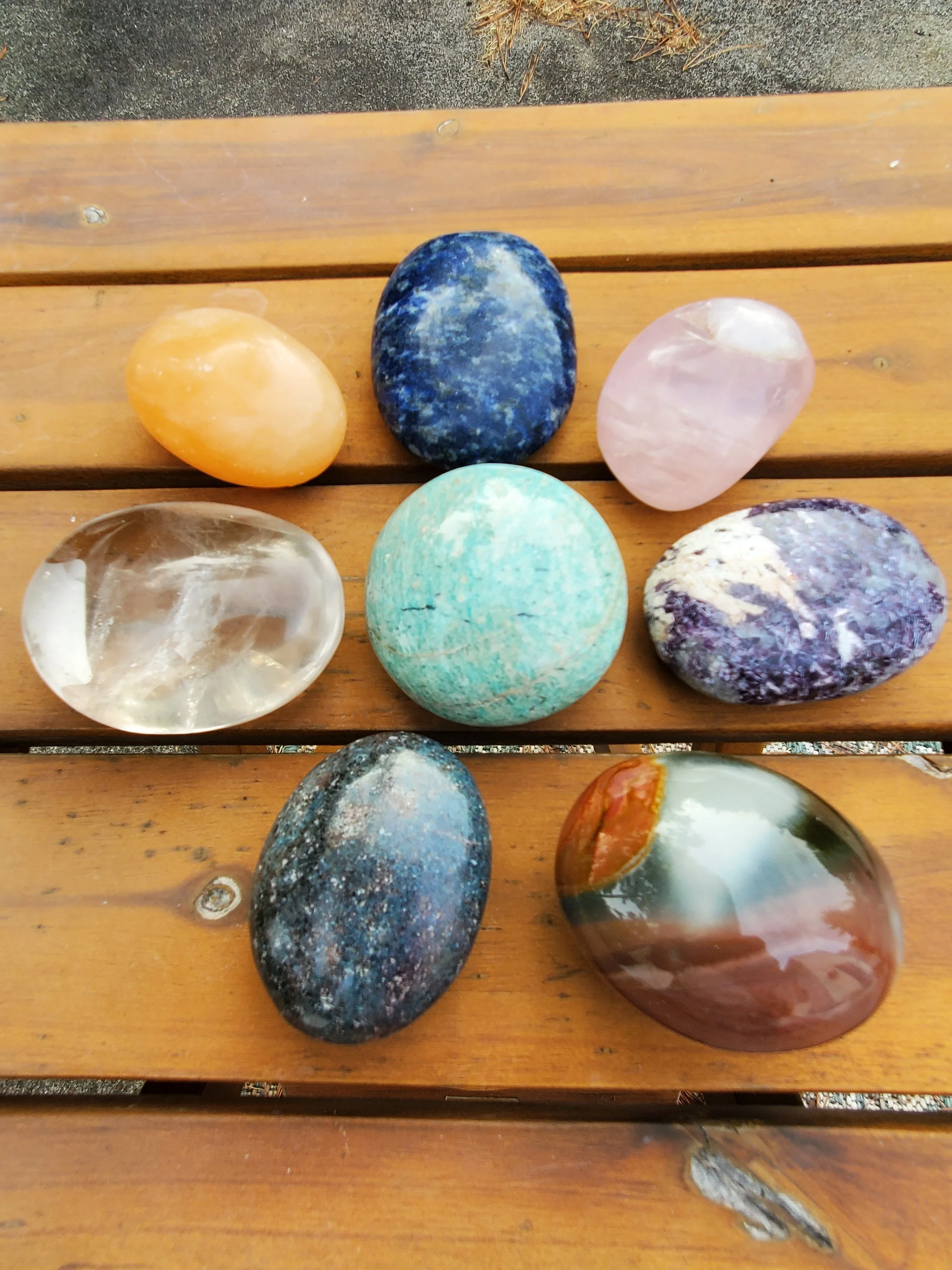 Nine polished stones of various colors and textures arranged on a wooden surface outdoors.