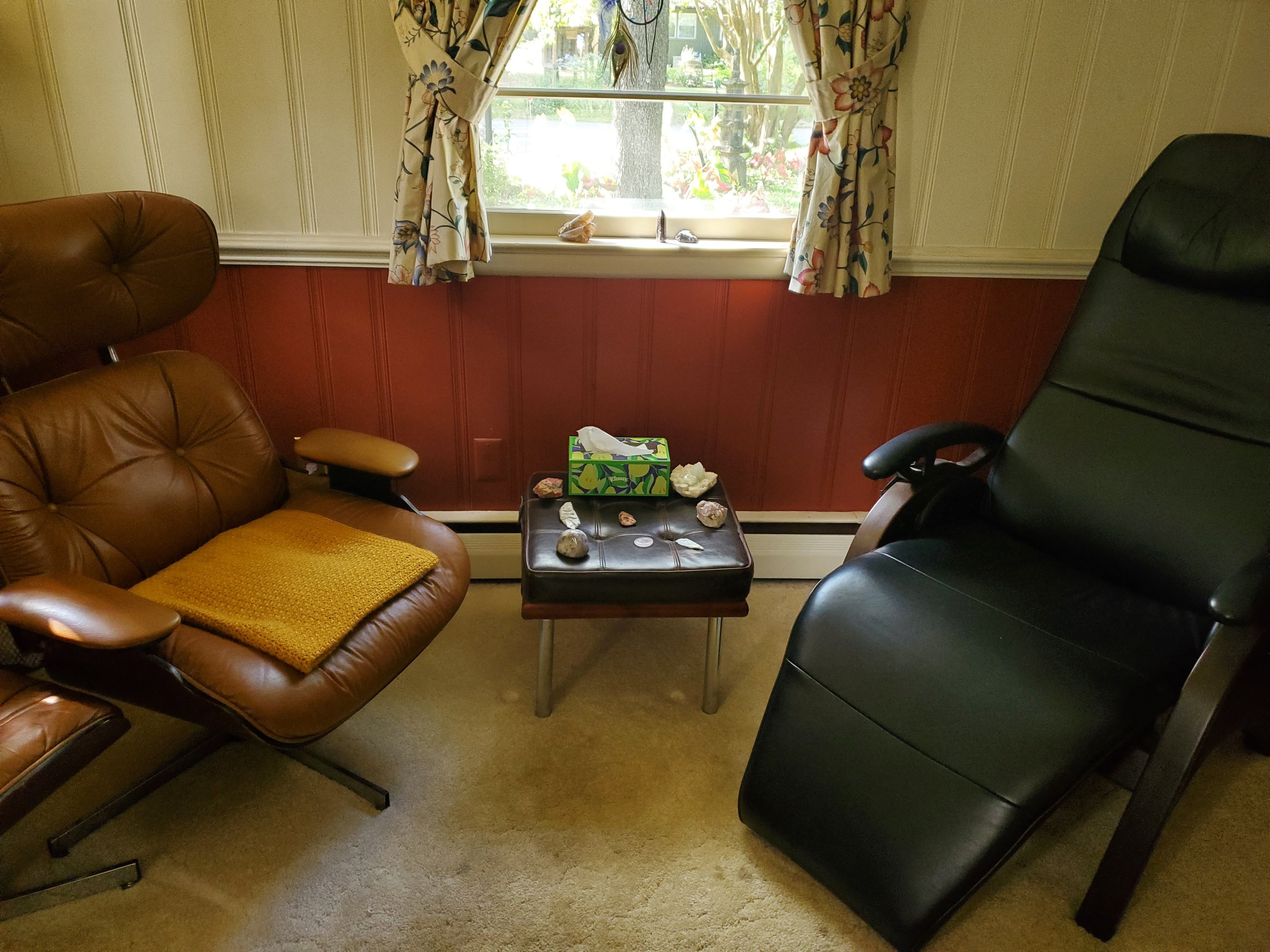 A cozy living room with a window above a red wood-paneled wall, featuring floral curtains. There is a brown leather chair with a yellow throw blanket on it and a black leather recliner. In between, a small black table holds a tissue box and several rocks.