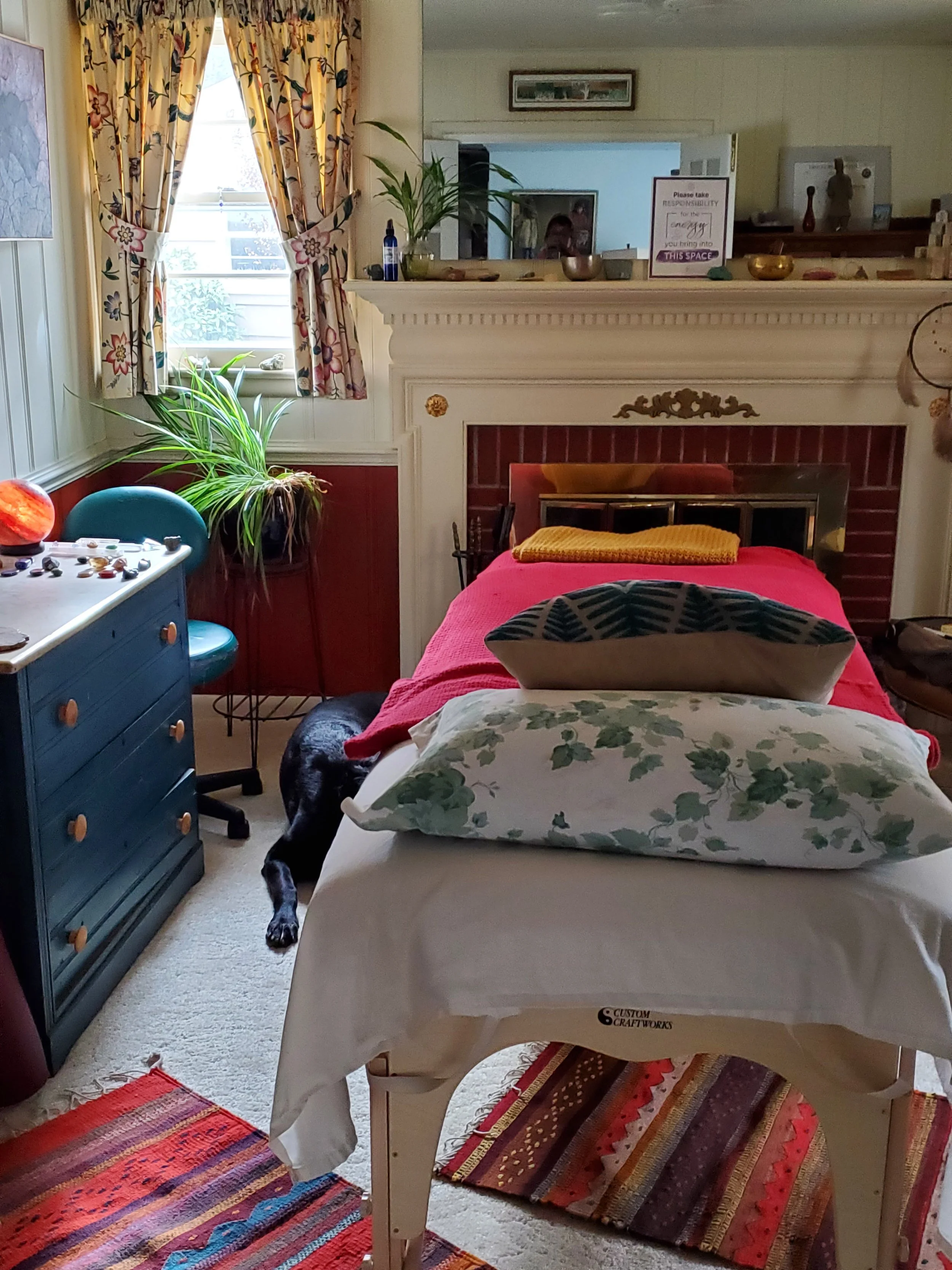 A cozy room with a window adorned with floral curtains, a fireplace with a mirror above, and a bed with colorful pillows and a red blanket. There is a blue dresser with a pumpkin on top, a green plant near the window, and a partially visible dog lying on the floor behind the bed. The room has various decorative items and rugs on the carpeted floor.