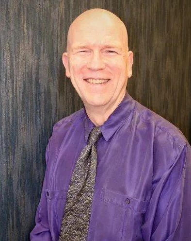 A smiling bald man wearing a purple dress shirt and a patterned tie, standing in front of a dark wooden background.