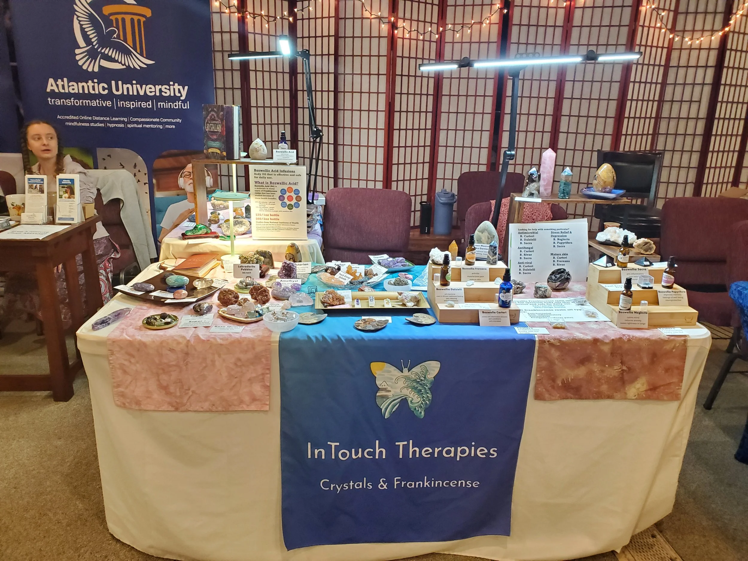 Table display of crystals, gemstones, and incense at an event, with informational signs, bottles, and decorative items. The table has a blue cloth with the logo and text 'InTouch Therapies Crystals & Frankincense.' In the background, there are chairs and a large Atlantic University banner.