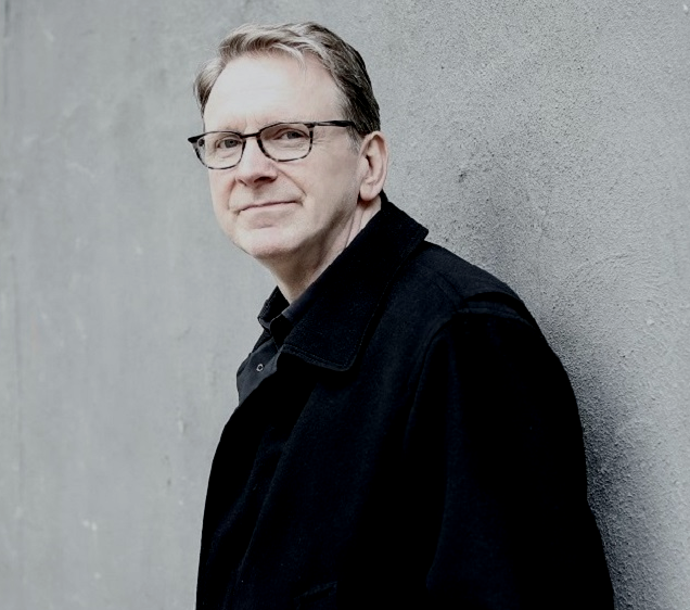 A middle-aged man with glasses and gray hair leaning against a gray concrete wall, wearing a black jacket and black shirt.