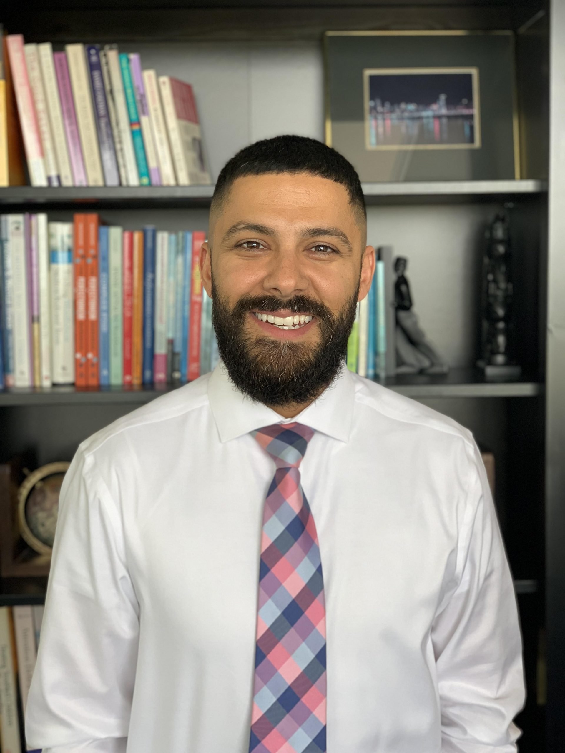 Smiling man in white shirt and multicolored tie standing in front of a bookshelf.
