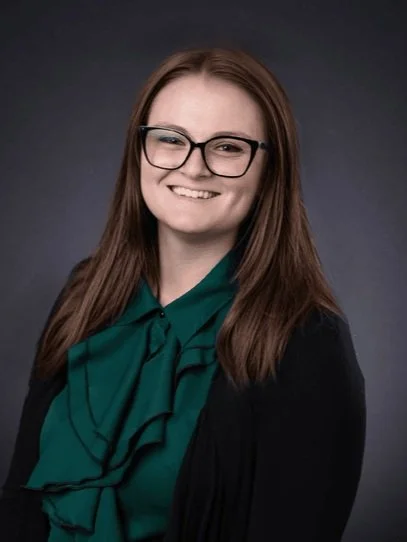 Person wearing glasses, with long brown hair, smiling, dressed in a green blouse and dark jacket, set against a gray background.