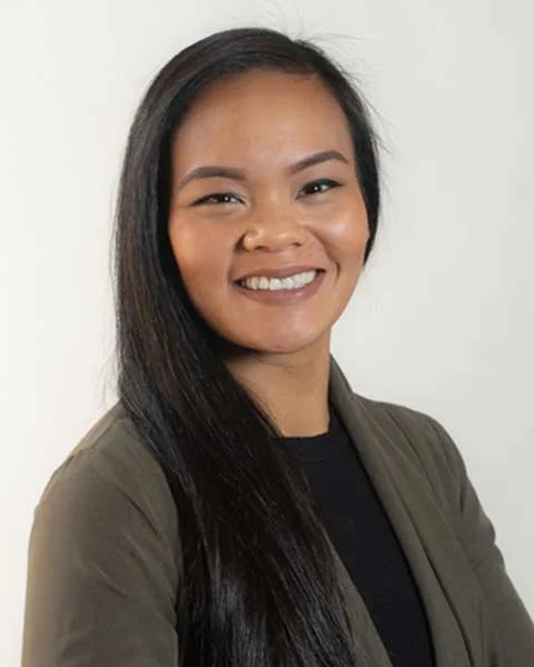 Smiling person with long hair, wearing a black shirt and gray blazer, on a light background.