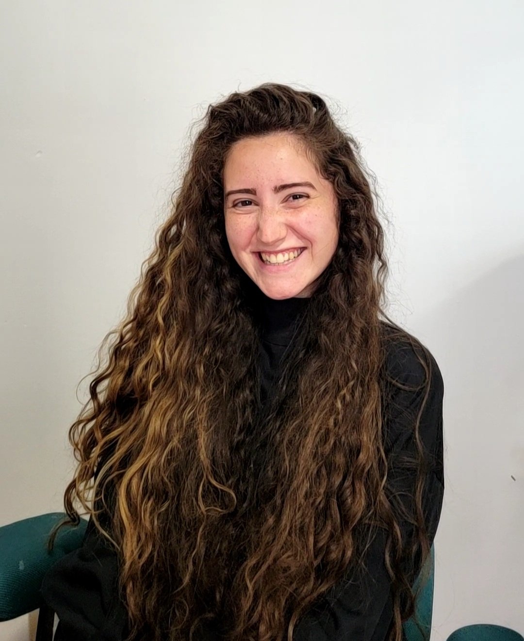 A person with long curly hair smiling at the camera, wearing a black top, seated on a green chair against a plain background.