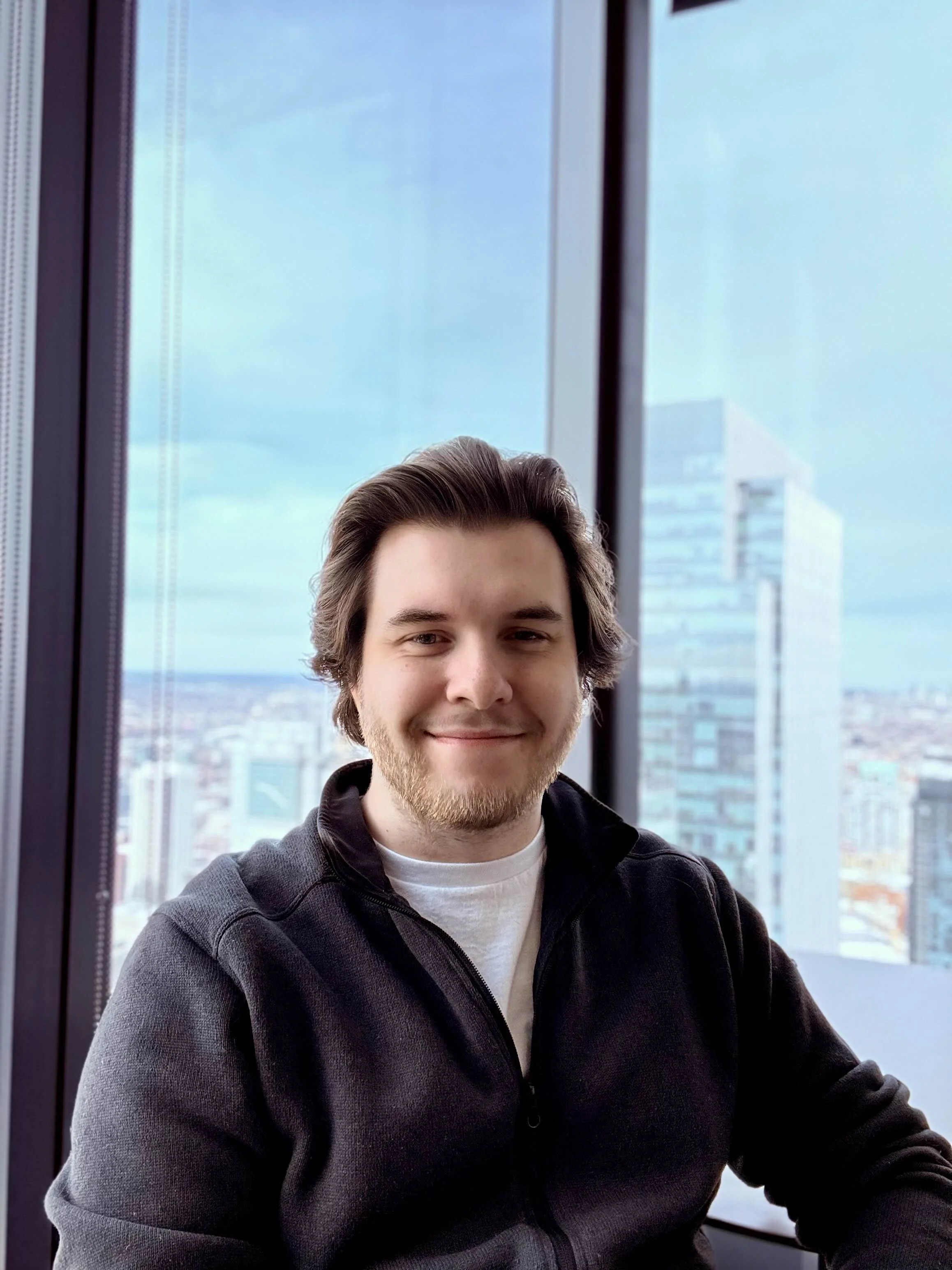 Person smiling indoors with cityscape background viewed through large windows.
