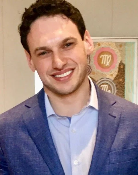 Man in blue blazer smiling, standing indoors with artistic background.
