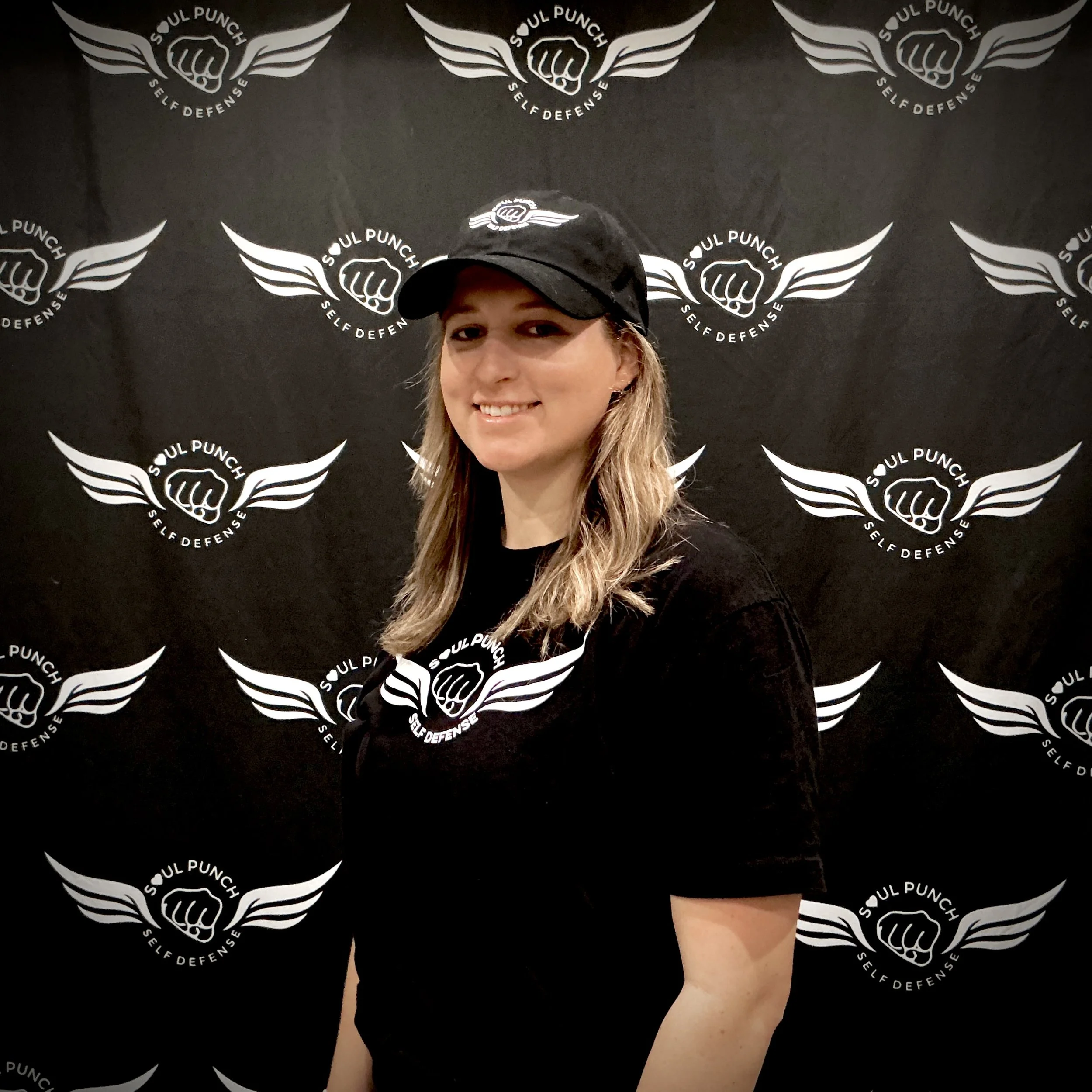 Photo of self-defense instructor Meg Wissman. Smiling young woman with long blonde hair wearing a black baseball cap and black t-shirt featuring the Soul Punch logo, standing in front of a black backdrop with the same logo repeated in a pattern.