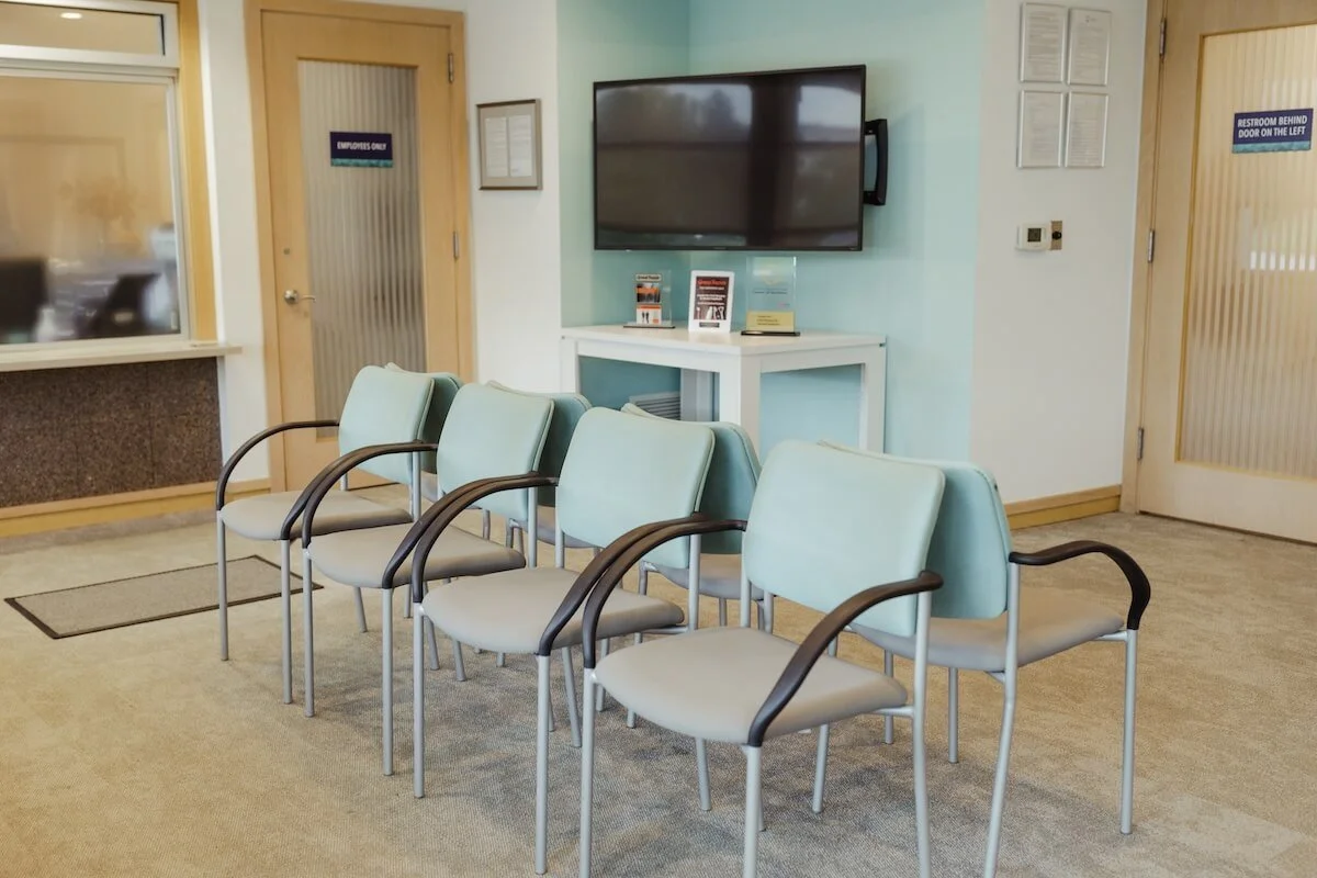 Empty waiting room with five light blue chairs with black armrests in front of a white table with brochures, a TV mounted on the wall, and two doors with blue signs indicating 'Employees Only' and 'Restroom Behind Door on the Left.'
