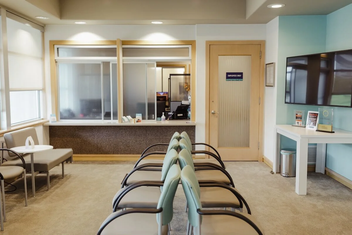 Waiting area in a medical office with chairs, a window, a front desk behind a glass partition, and a TV on the wall.