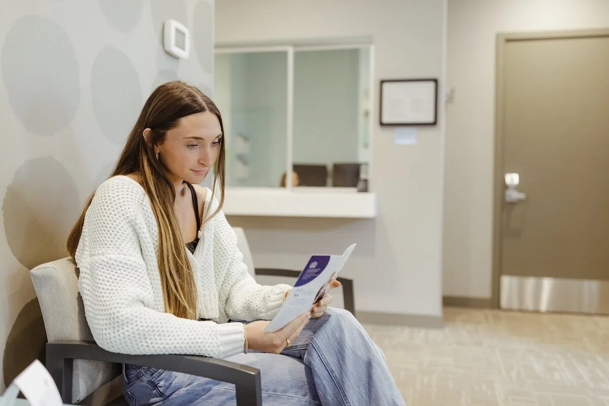 A young woman sitting in a waiting room, reading a brochure or pamphlet.
