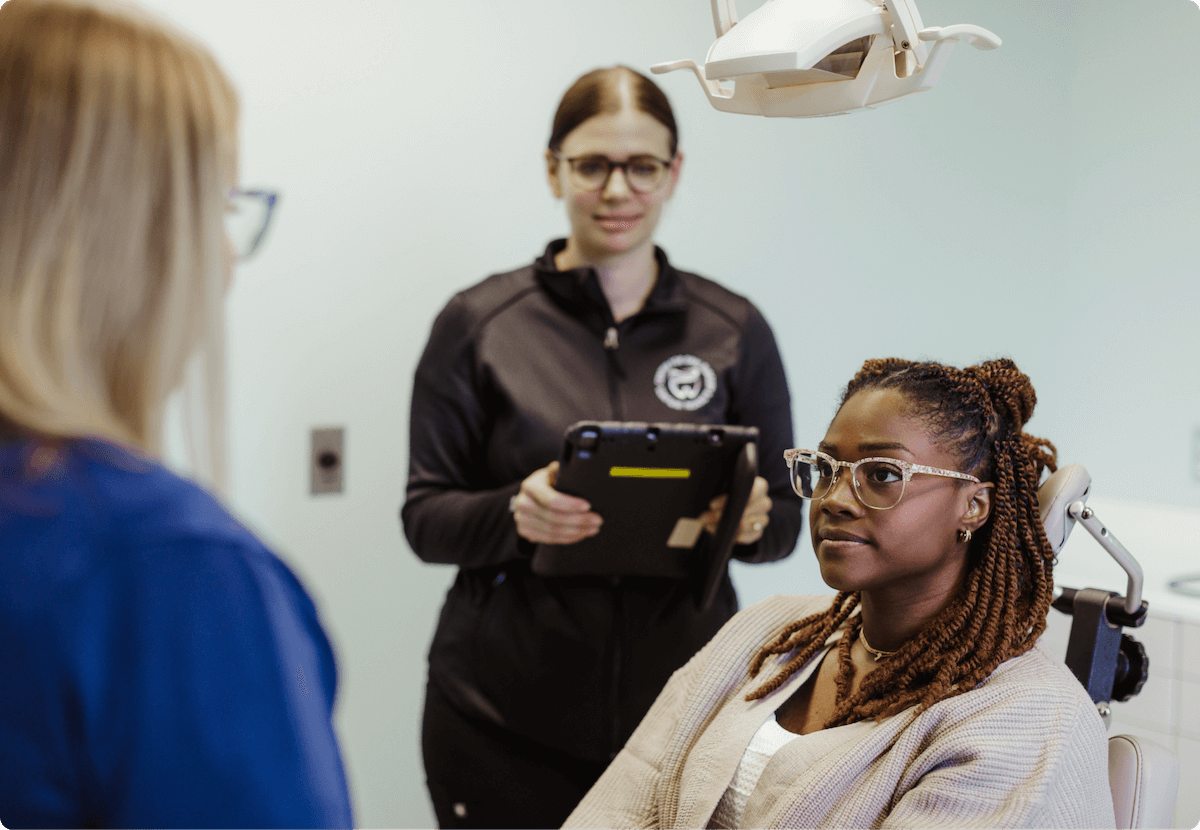 A young woman with glasses and braids sitting in a dental chair, talking to a woman in blue medical scrubs, with a dental light overhead and a woman with glasses and a black jacket standing nearby holding a tablet.