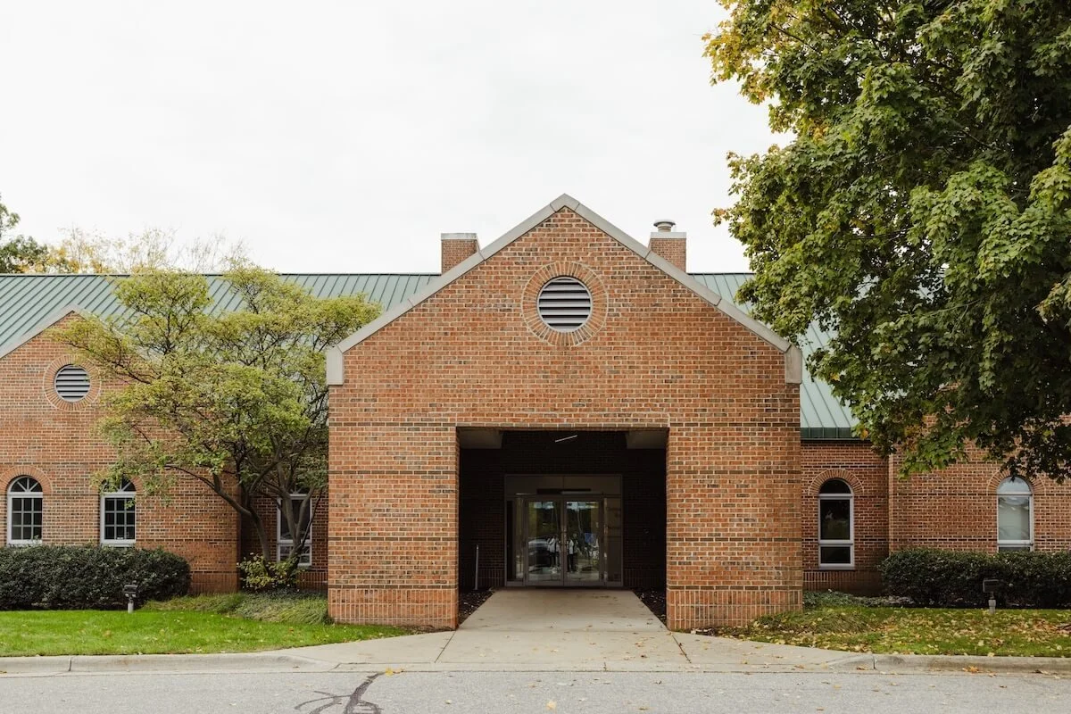 Red brick building with a centered entrance, flanked by trees and shrubs, and a green metal roof.