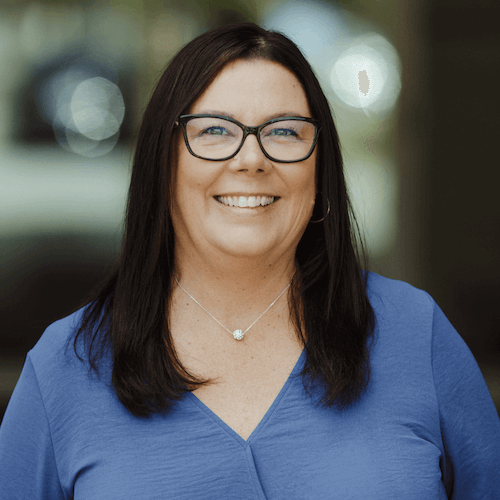 Portrait of a woman with long dark hair, glasses, and a blue V-neck top, smiling outdoors