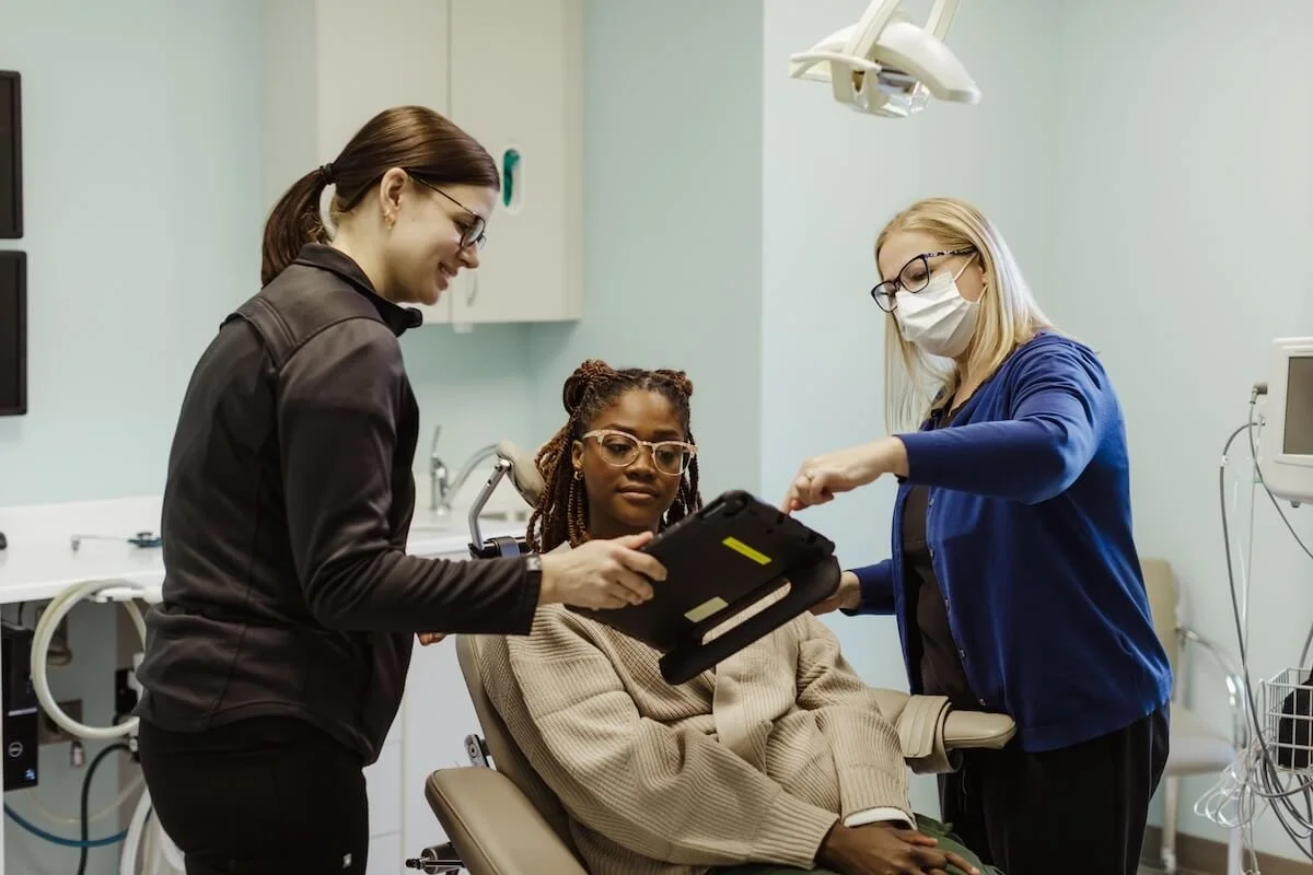 Two female healthcare professionals and a woman patient in a medical examination room, with the professionals discussing medical equipment.