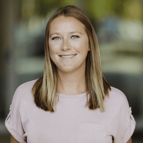 A woman with shoulder-length light brown hair, smiling, wearing a light pink t-shirt, standing outdoors with blurred trees in the background.